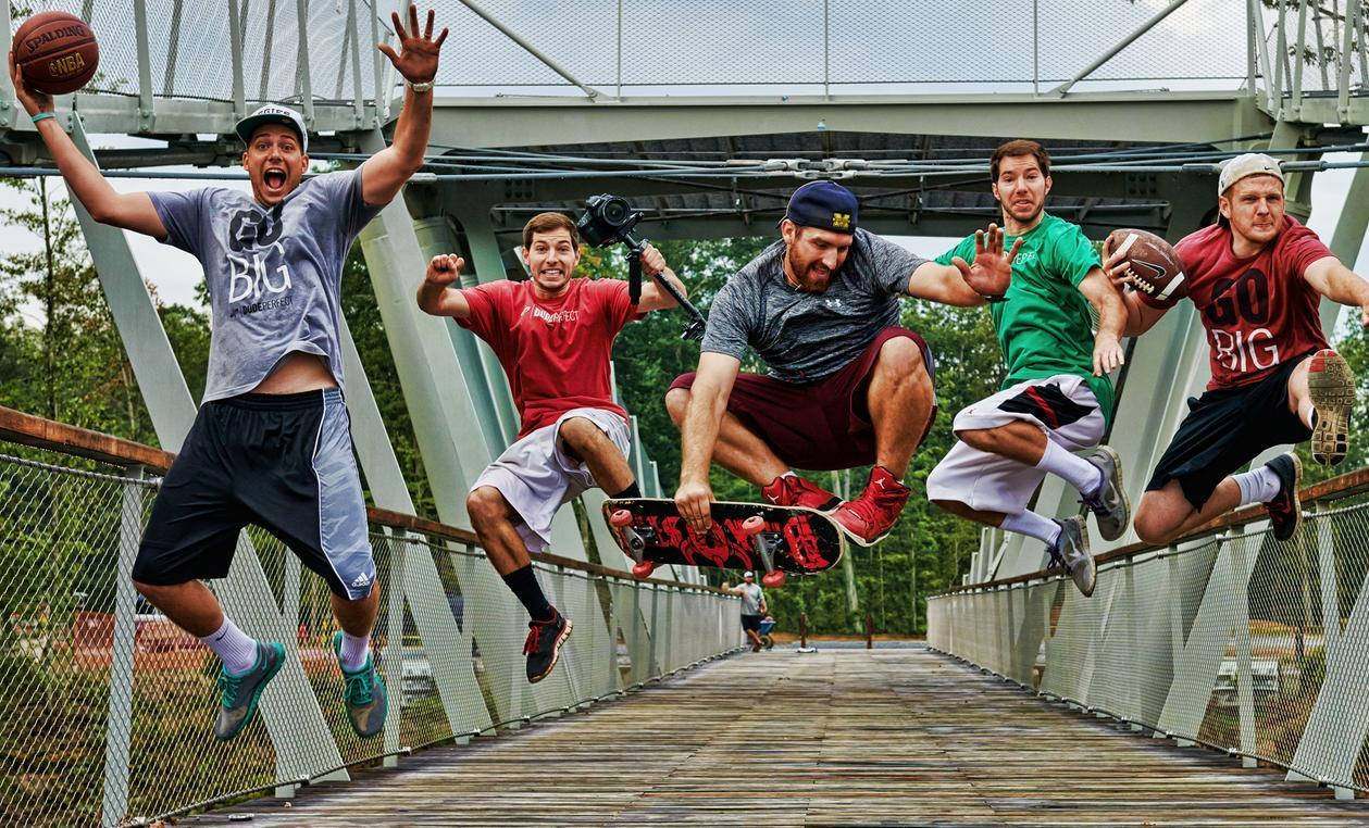 Dude Perfect 團隊從橋上跳水 壁紙 — 五個男人喺行人天橋上半空，一個拎住籃球，另一個拎住足球，一個踩緊滑板，全部定格喺充滿活力嘅動作中。