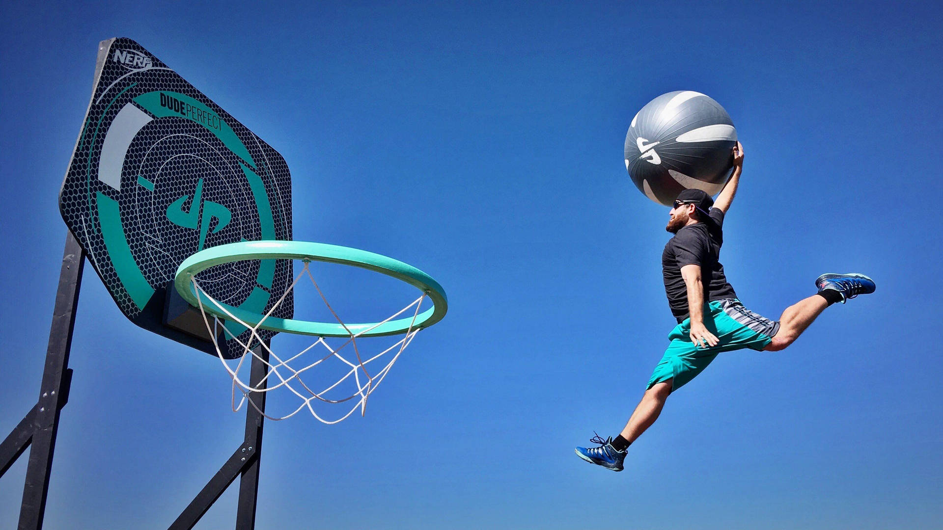 Dude Perfect 超大號籃球框 壁紙 — 一名男子在晴朗的藍天下，於青綠色籃球框附近高舉一個大型健身球跳起。