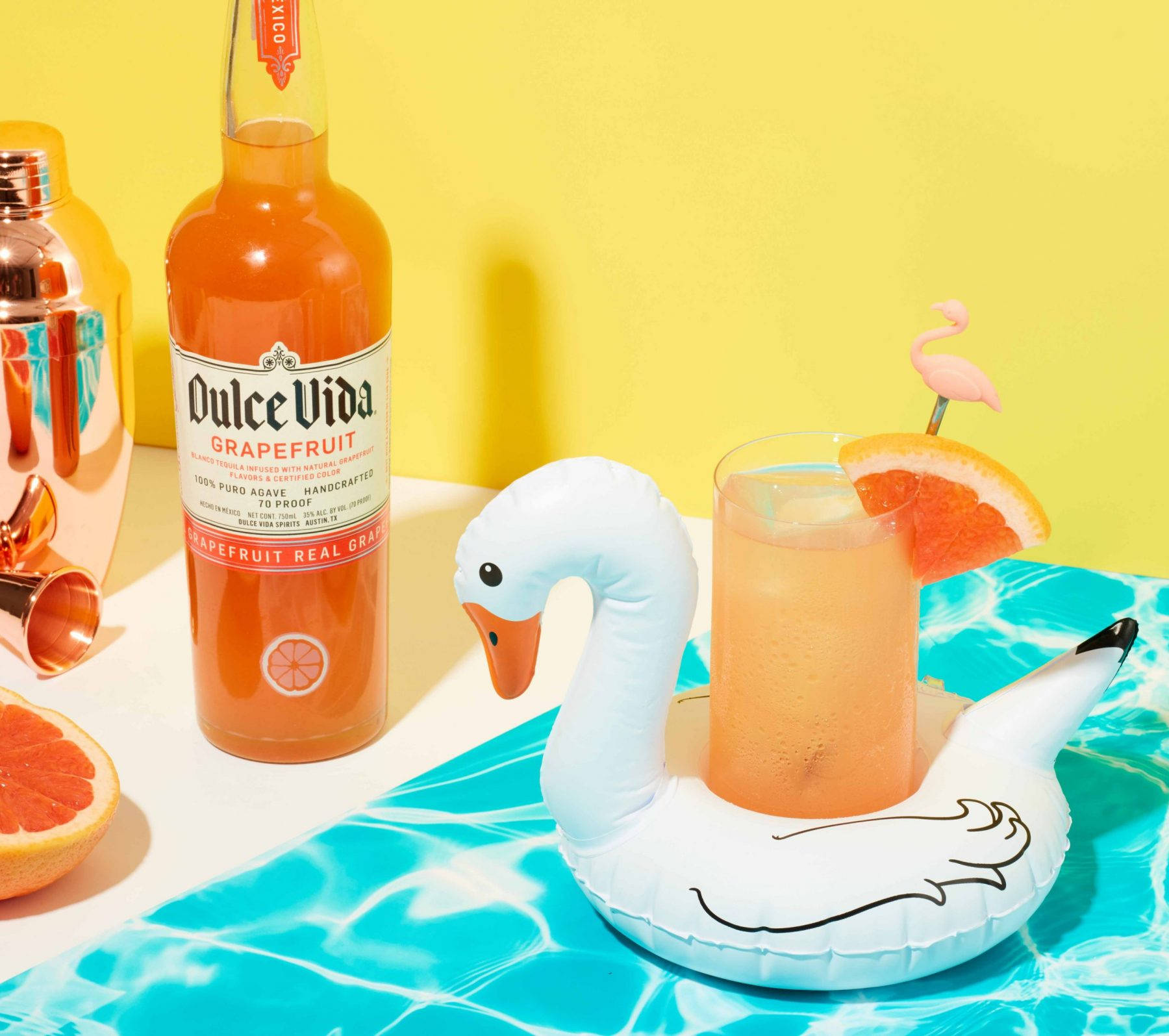 夏日派對雞尾酒 Dulce Vida 飲品 壁紙 — 在陽光普照的黃色牆壁背景下，泳池邊桌上有一個天鵝浮圈，旁邊放著一瓶西柚，裡面盛著充滿活力的熱帶飲品。