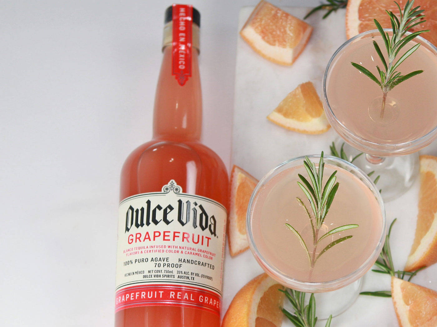 Dulce Vida 風味烈酒派對飲品 壁紙 — 一個充滿活力的粉紅西柚龍舌蘭酒瓶旁邊有兩杯以迷迭香和柑橘片裝飾的雞尾酒，放在白色大理石表面上。