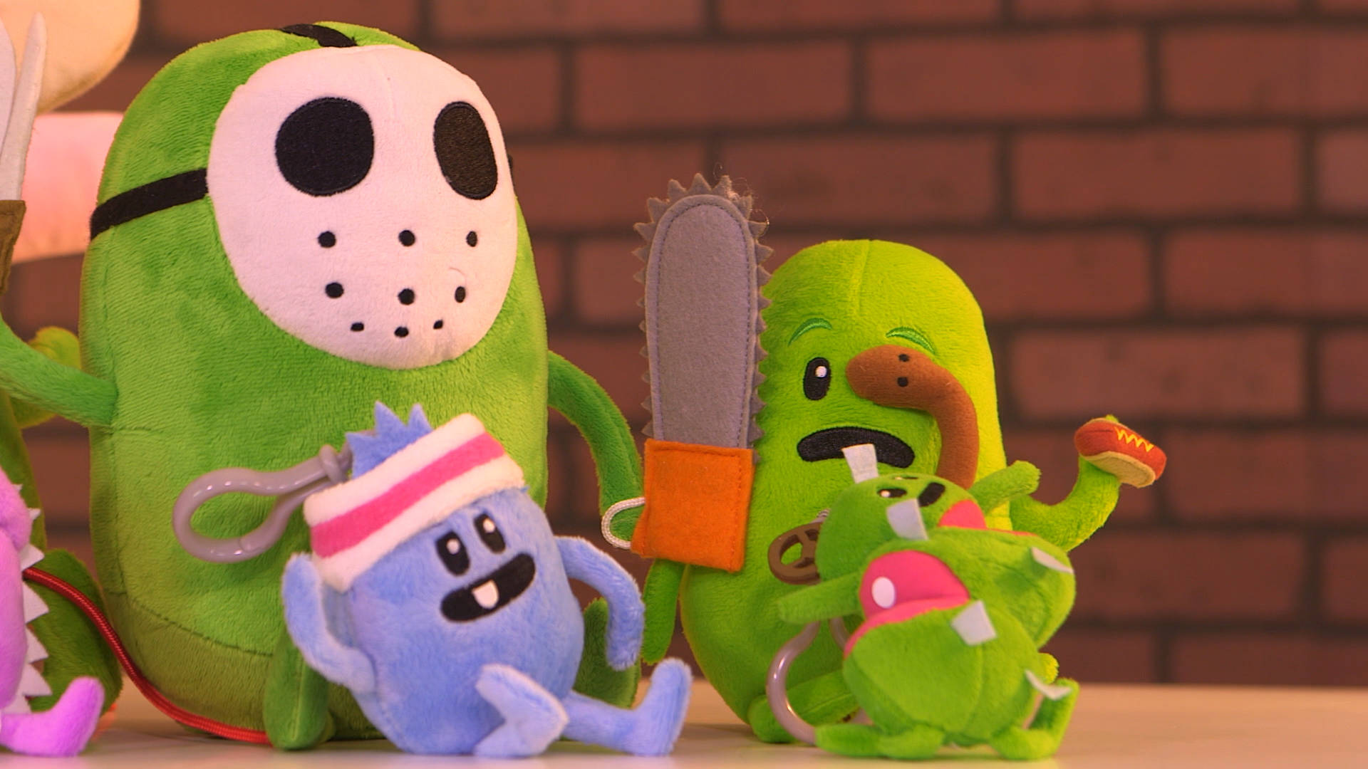 Dumb Ways To Die 毛絨玩具版 壁紙 — 綠色和藍色卡通角色的毛絨玩具，帶著電鋸和曲棍球面具，在磚牆前擺姿勢。