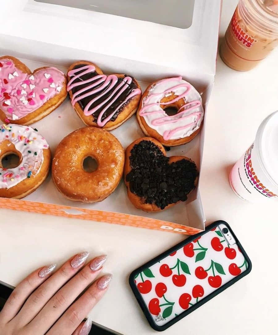 享用Dunkin' Donuts香醇冰萃咖啡 壁紙 — 從上方俯瞰一個裝滿心形和糖霜甜甜圈的Dunkin' Donuts盒子，旁邊有櫻桃圖案的手機殼和冰咖啡。