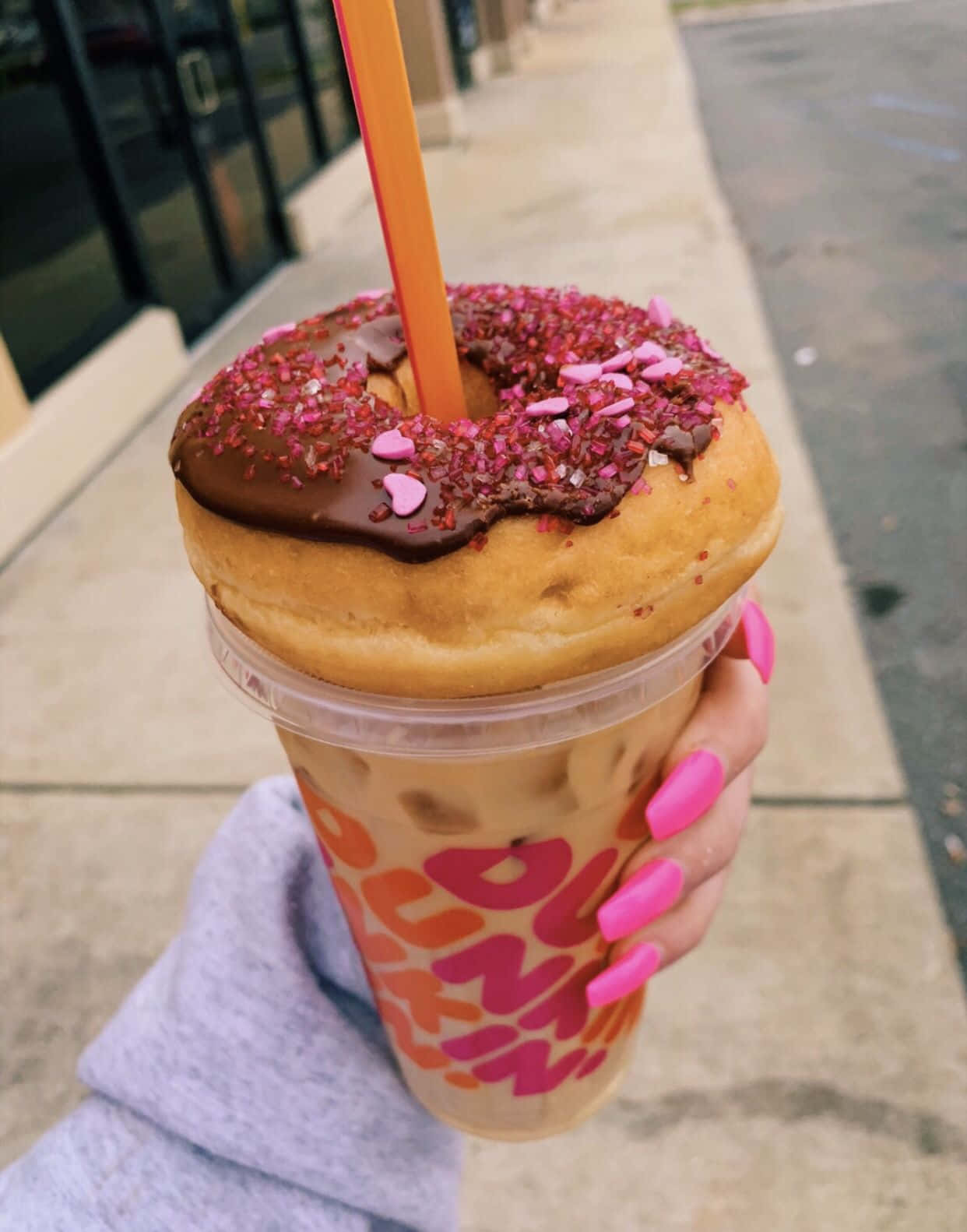 Dunkin' Donuts新鮮沖煮咖啡與美味甜甜圈 壁紙 — 一隻手在人行道上拿著一杯Dunkin'冰咖啡，上面有一個朱古力釉面冬甩和粉紅色糖針。
