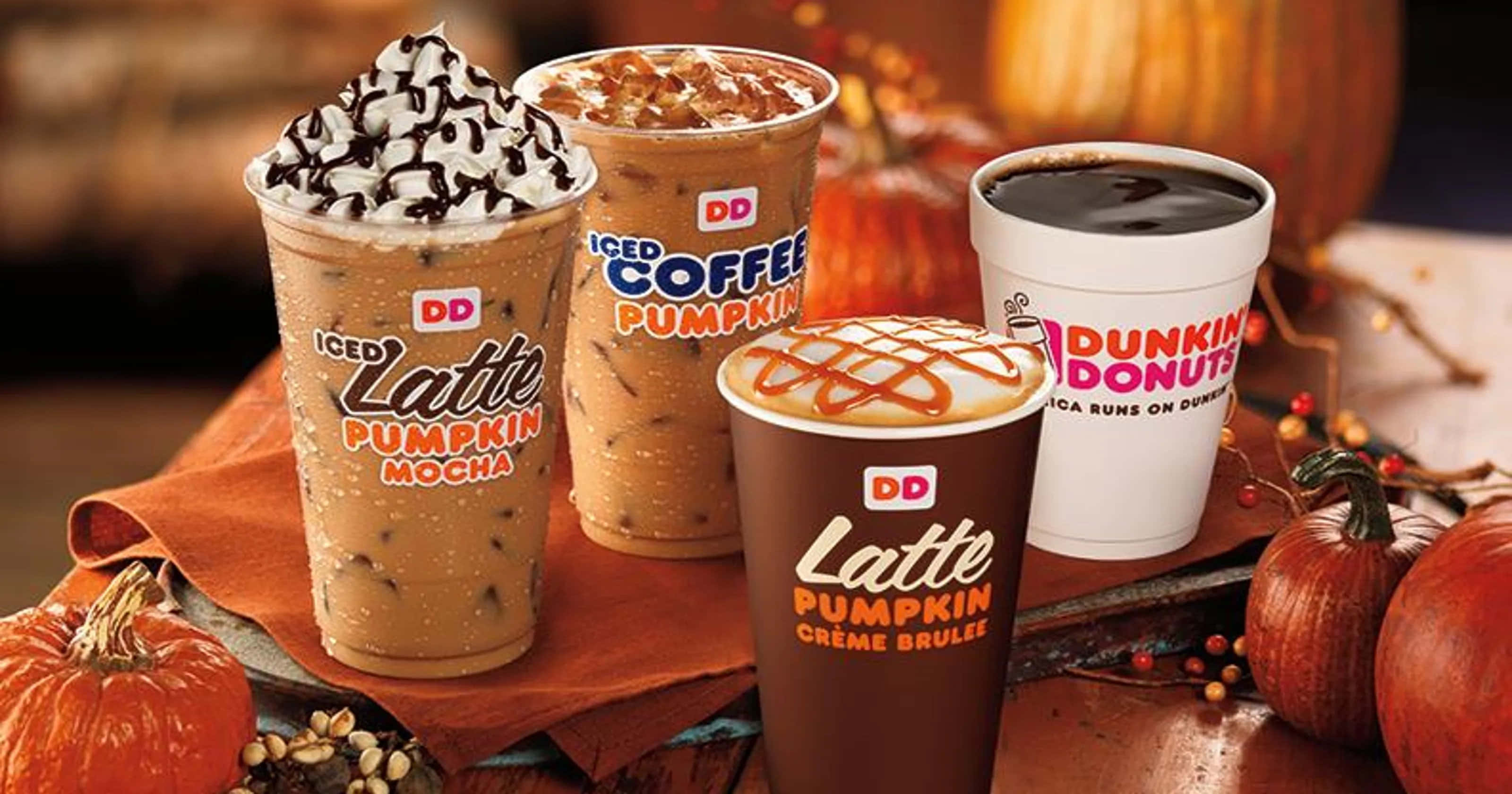 以Dunkin' Donuts開啟美好一天 壁紙 — 四杯Dunkin' Donuts南瓜味飲品，上面有忌廉、焦糖和朱古力醬，周圍有南瓜和秋季裝飾。