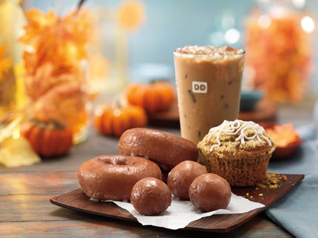 滿足您的甜蜜渴望，盡享Dunkin' Donuts 壁紙 — 一個木托盤上放有釉面甜甜圈、朱古力松露和南瓜香料鬆餅，旁邊是一杯冰咖啡，背景中有秋葉和南瓜，呈現柔和焦點。