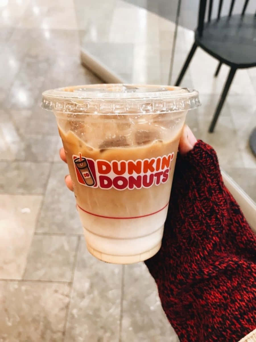 以您最愛的Dunkin' Donuts風味醒來 壁紙 — 一隻穿著紅色針織袖子的手，拿著一杯有冰塊的Dunkin' Donuts凍咖啡，背景是瓷磚地板和椅子。