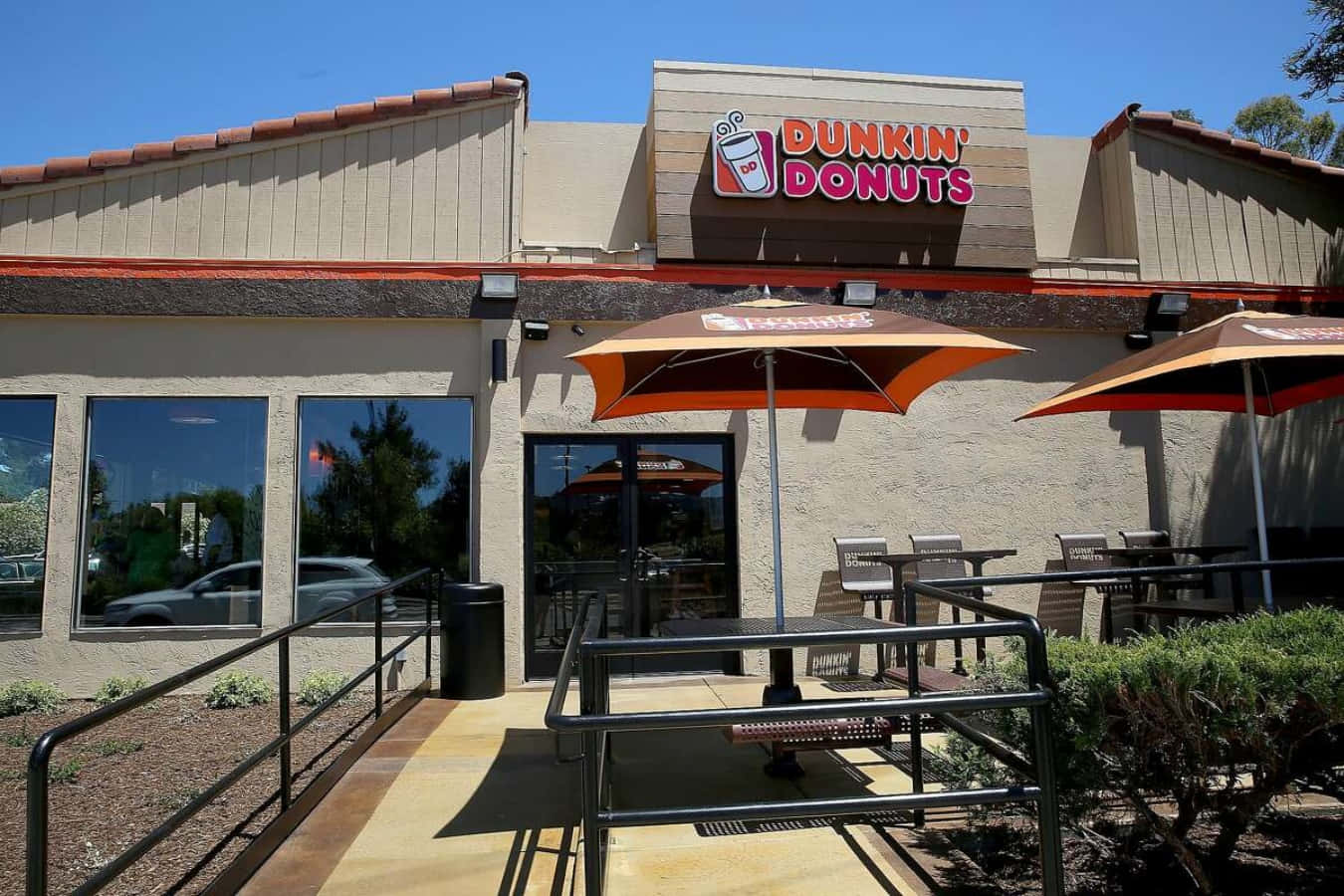 以一杯咖啡和甜甜圈開始美好的一天，來自Dunkin' Donuts！ 壁紙 — Dunkin' Donuts 店舖前一個晴朗的日子，橙色遮陽傘為戶外座位提供陰涼，上方是清澈的藍天。