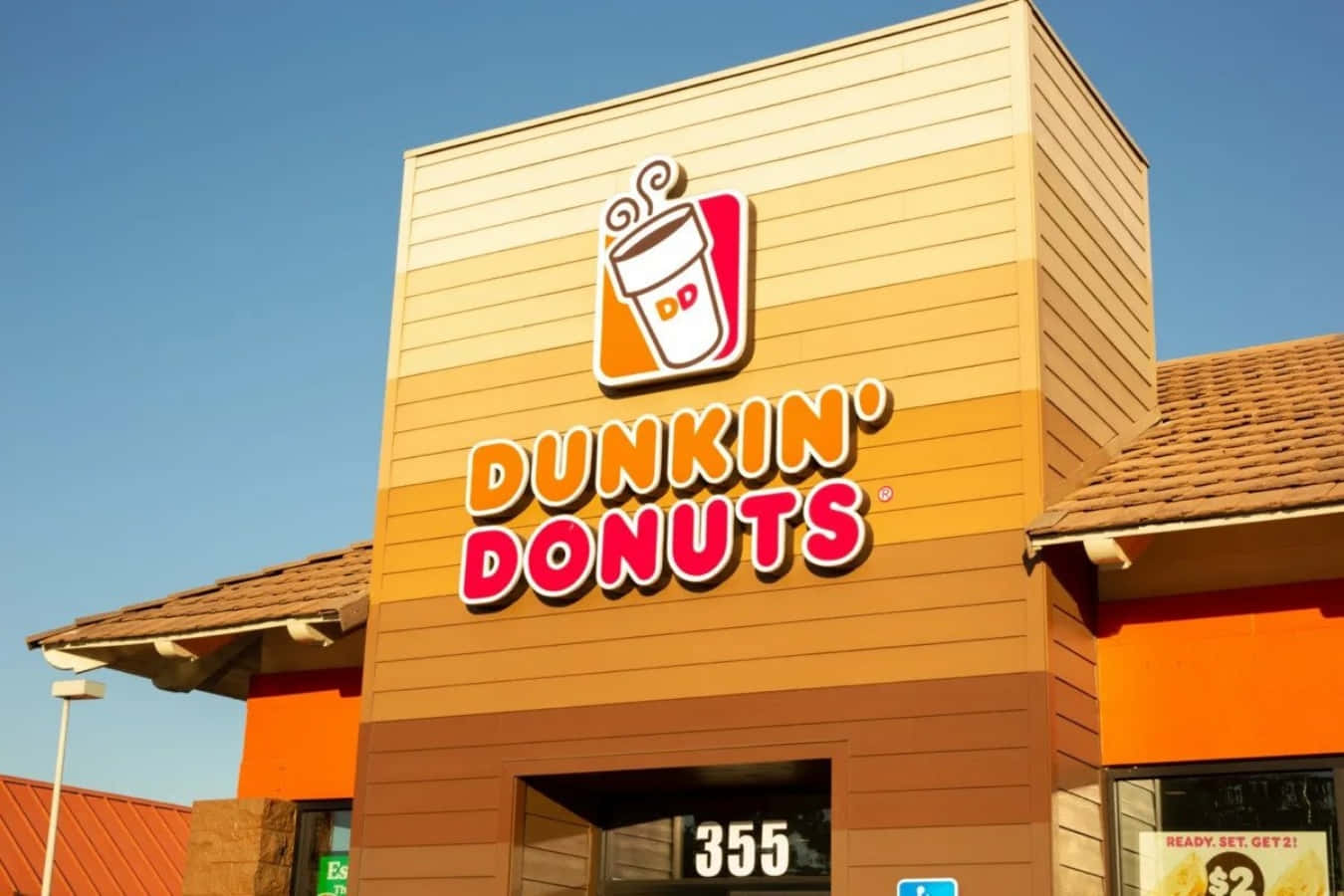 享受經典Dunkin' Donuts咖啡 壁紙 — 晴朗藍天下，明亮的橙色Dunkin' Donuts店面，配以醒目的招牌和溫暖的陽光。