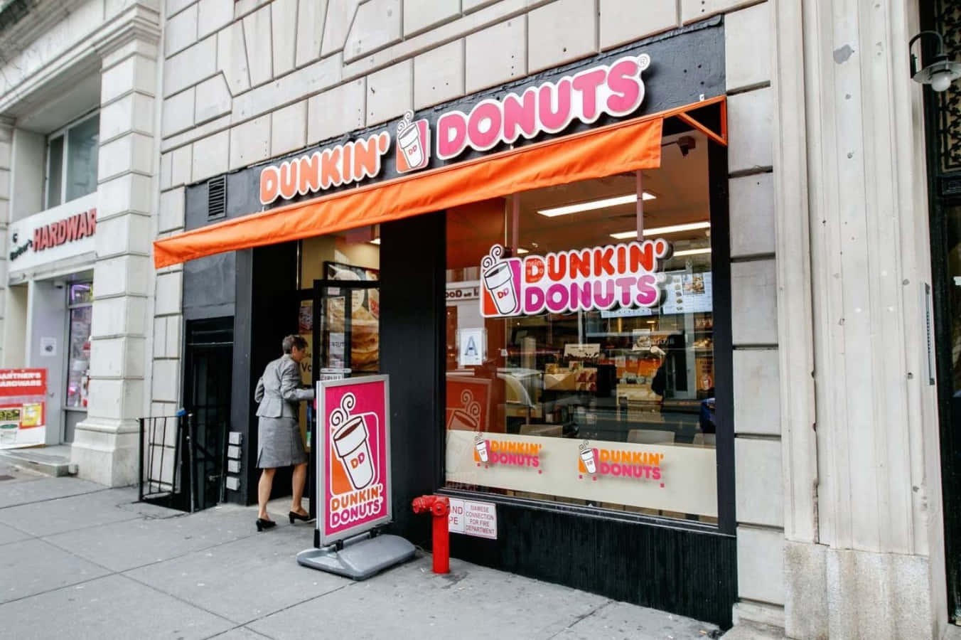 盡情享受Dunkin' Donuts的美味 壁紙 — 一位身穿灰色西裝的女士在城市行人路上走過一間Dunkin' Donuts店舖前鮮橙色的簷篷。