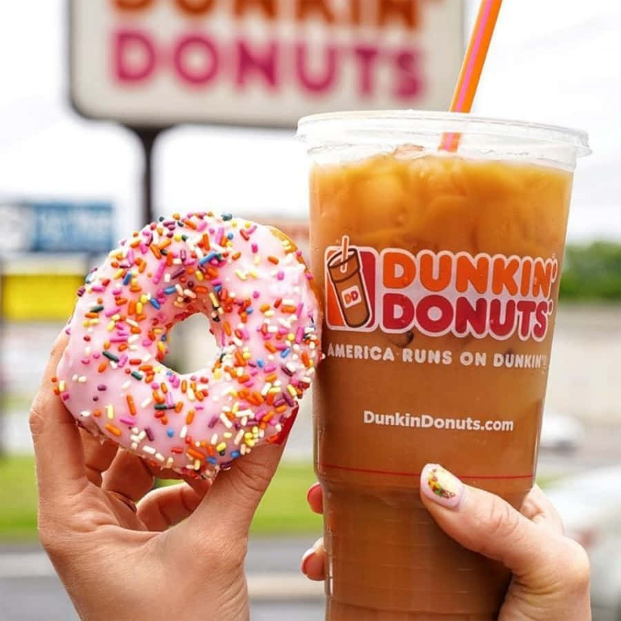 渴望Dunkin' Donuts的絕妙風味 壁紙 — 一隻手拿著一個粉紅色糖霜彩針甜甜圈，旁邊放著一杯附有吸管的Dunkin' Donuts凍咖啡。