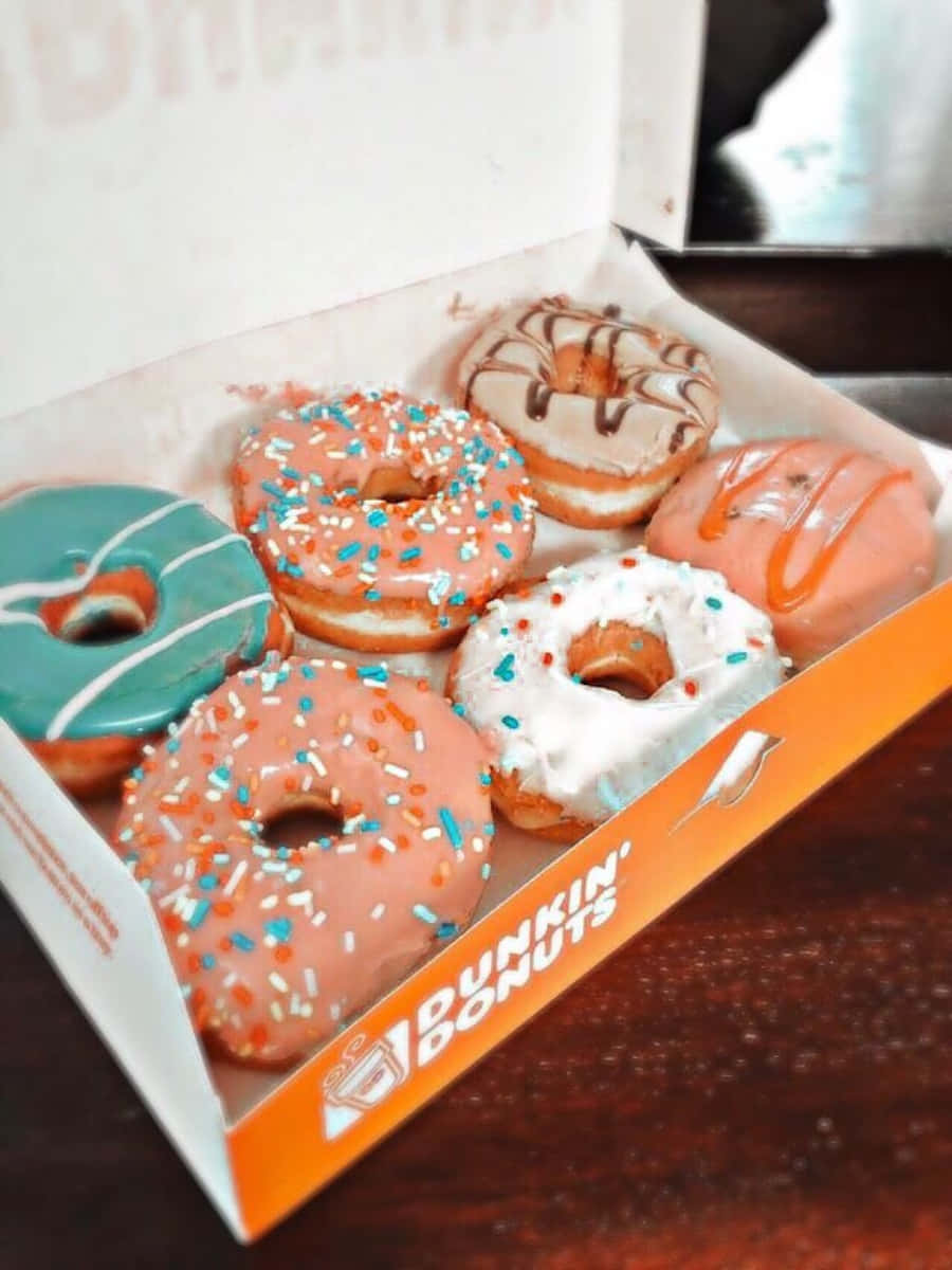 品嚐Dunkin' Donuts經典甜甜圈 壁紙 — 一盒色彩繽紛的Dunkin' Donuts冬甩，有粉紅、藍和白色糖霜及彩針，放在木桌上。