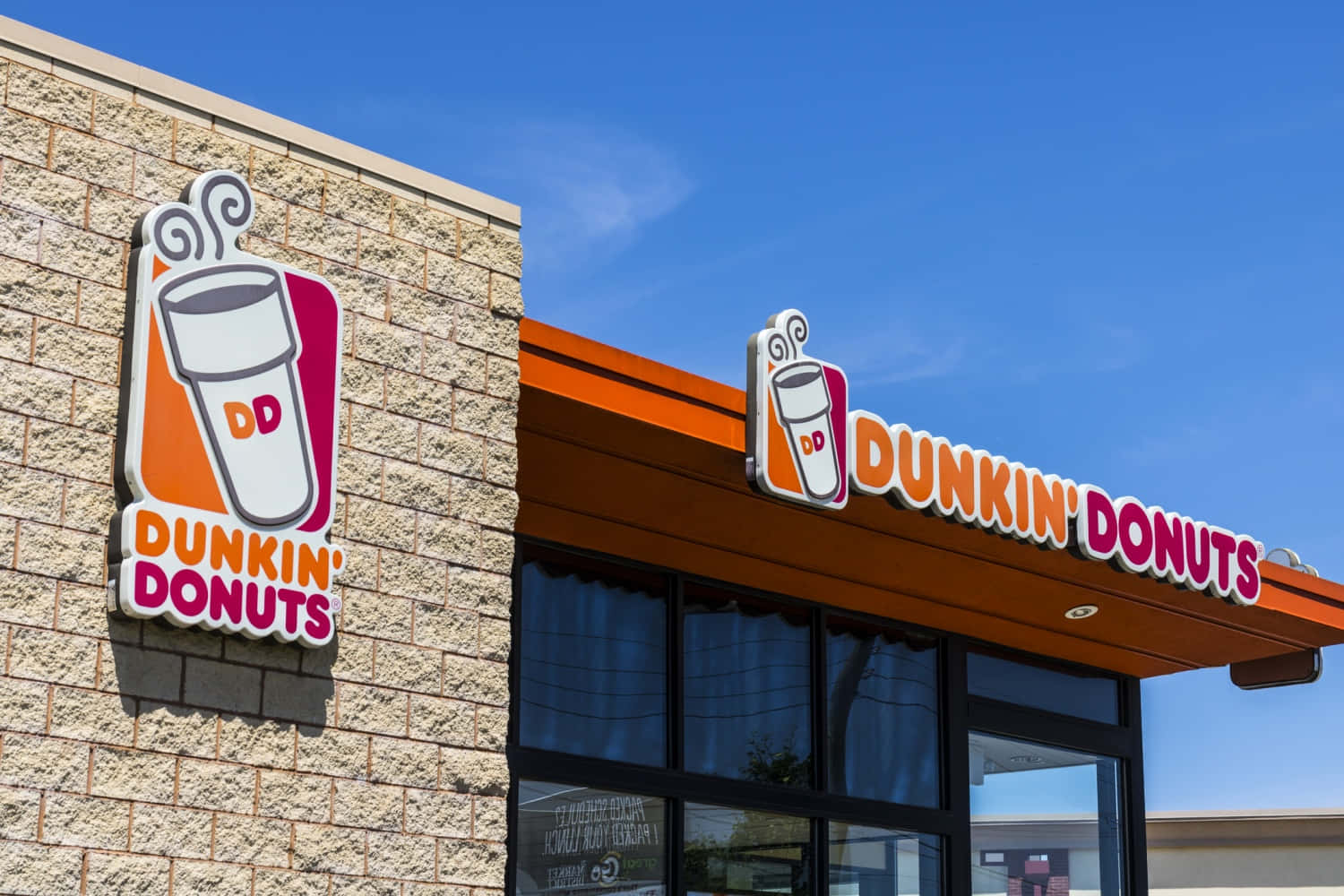 Dunkin' Donuts的歡樂時光 壁紙 — 明亮的藍天襯托著Dunkin' Donuts的店面，店面有醒目的橙色招牌和石磚牆。