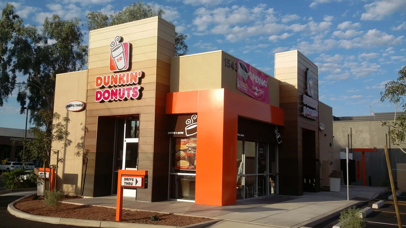 Dunkin' Donuts 的清新早晨享受 壁紙 — 在部分多雲的藍天下，一間亮橙色和米色的Dunkin' Donuts店舖外觀，有樹木和得來速標誌。