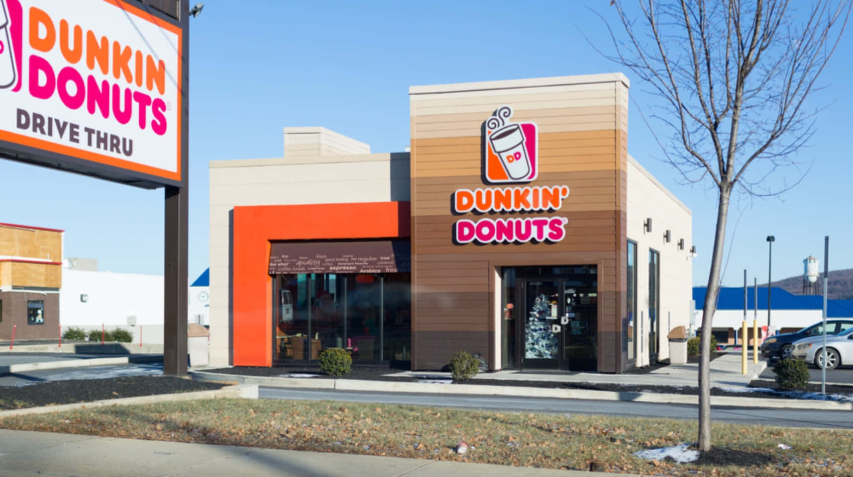 Dunkin' Donuts咖啡與甜點 壁紙 — 在晴朗藍天下，明亮的橙色和米色Dunkin' Donuts店面，有光禿禿的樹木和停放的汽車。