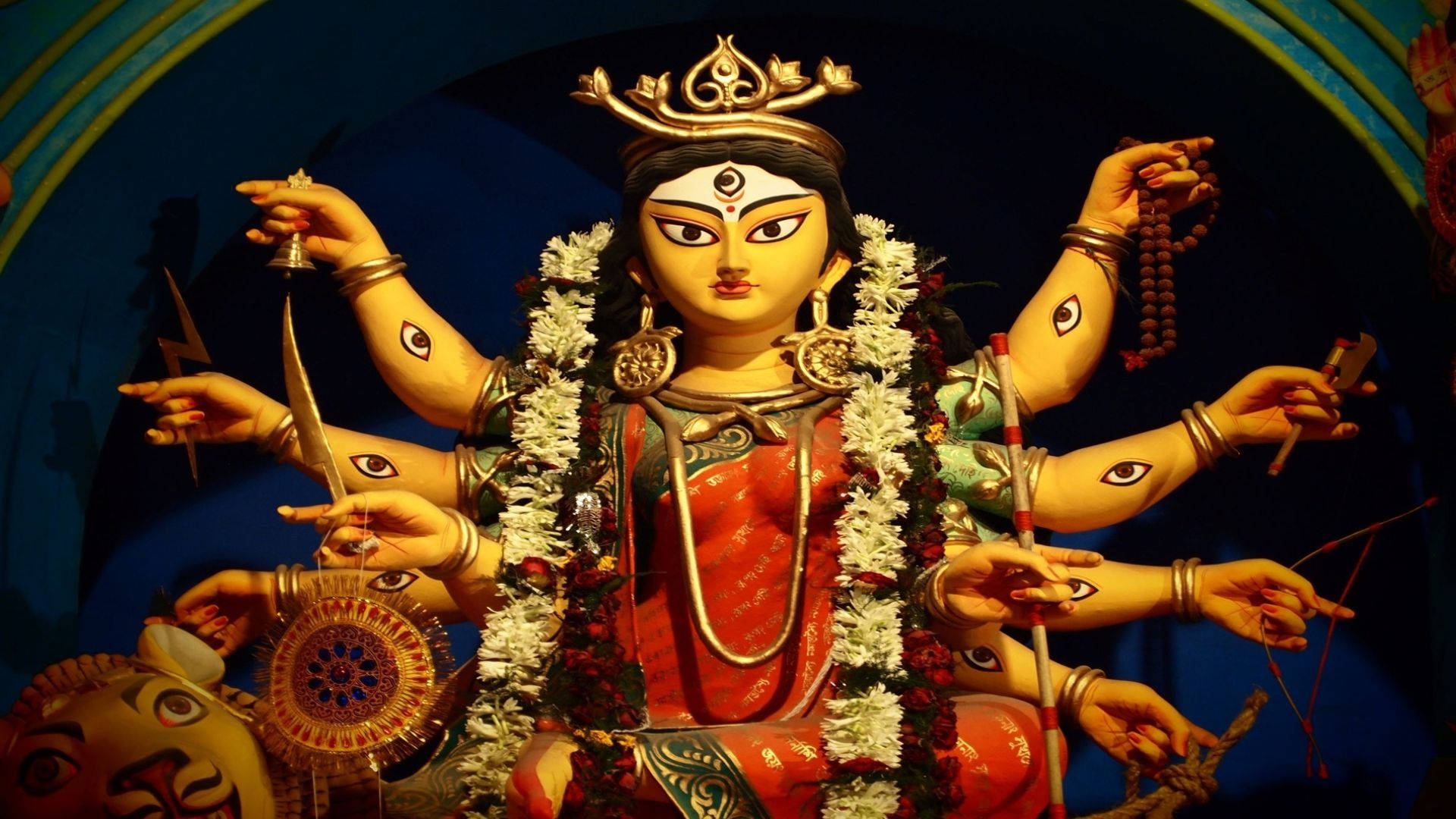 Durga Mata 高清雙眼 壁紙 — 一尊色彩鮮豔、多臂的印度教女神雕像，飾有白花、金飾和紅衣，在戲劇性的藍色燈光下。