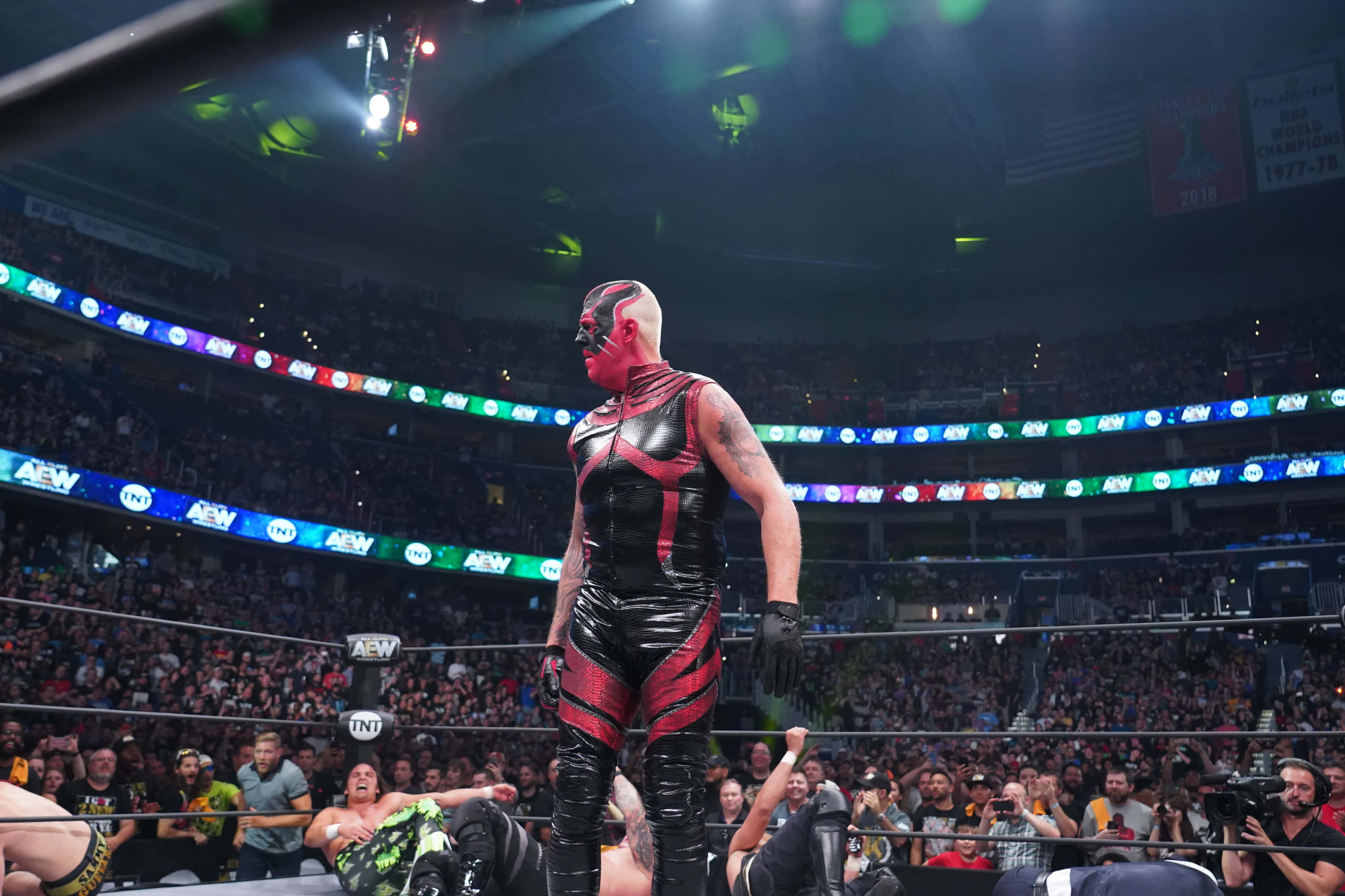 Dustin Rhodes 與 Britt Baker 對決 壁紙 — 一位戴面具的摔角手身穿紅黑戰衣，在明亮的場館燈光下站在擂台上，台下觀眾歡呼雷動。