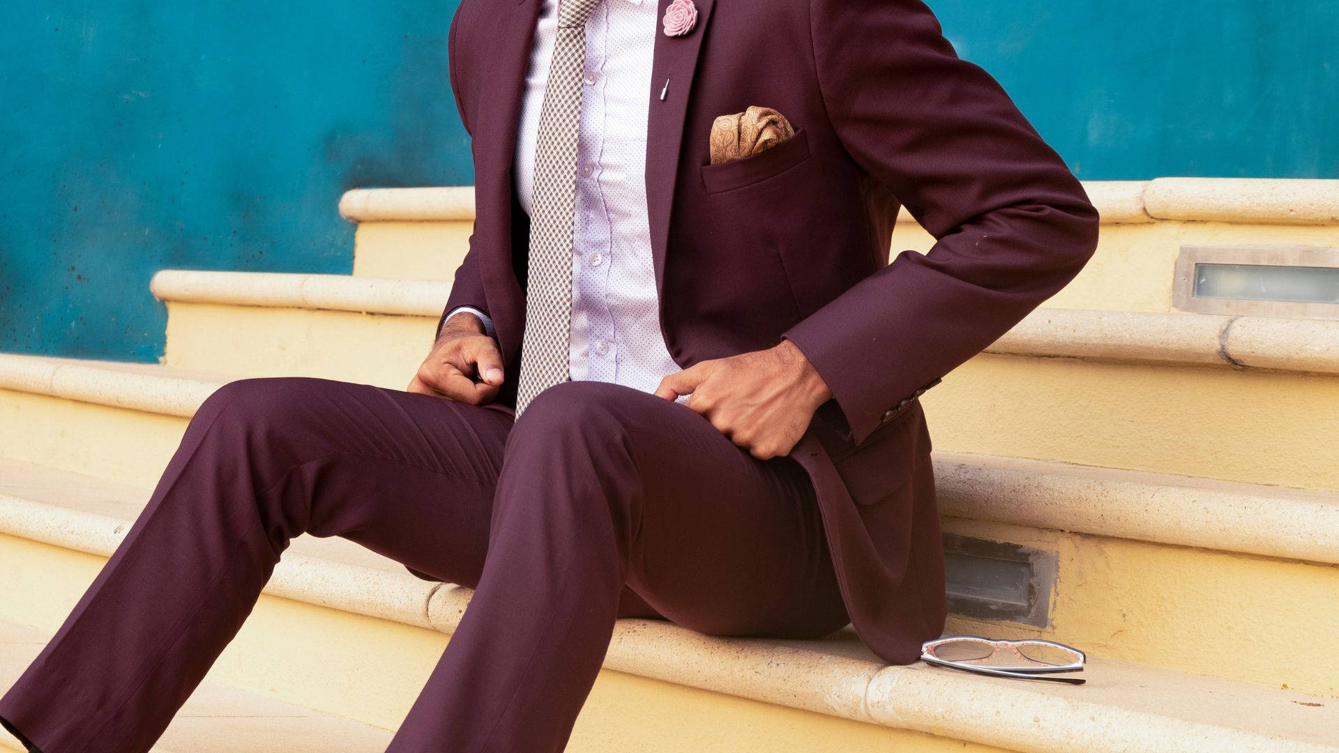 Dutch Menswear 定製剪裁西裝 壁紙 — 一位穿著棗紅色西裝的時尚男士自信地坐在黃色階梯上，背景是藍綠色牆壁，太陽眼鏡放在旁邊。