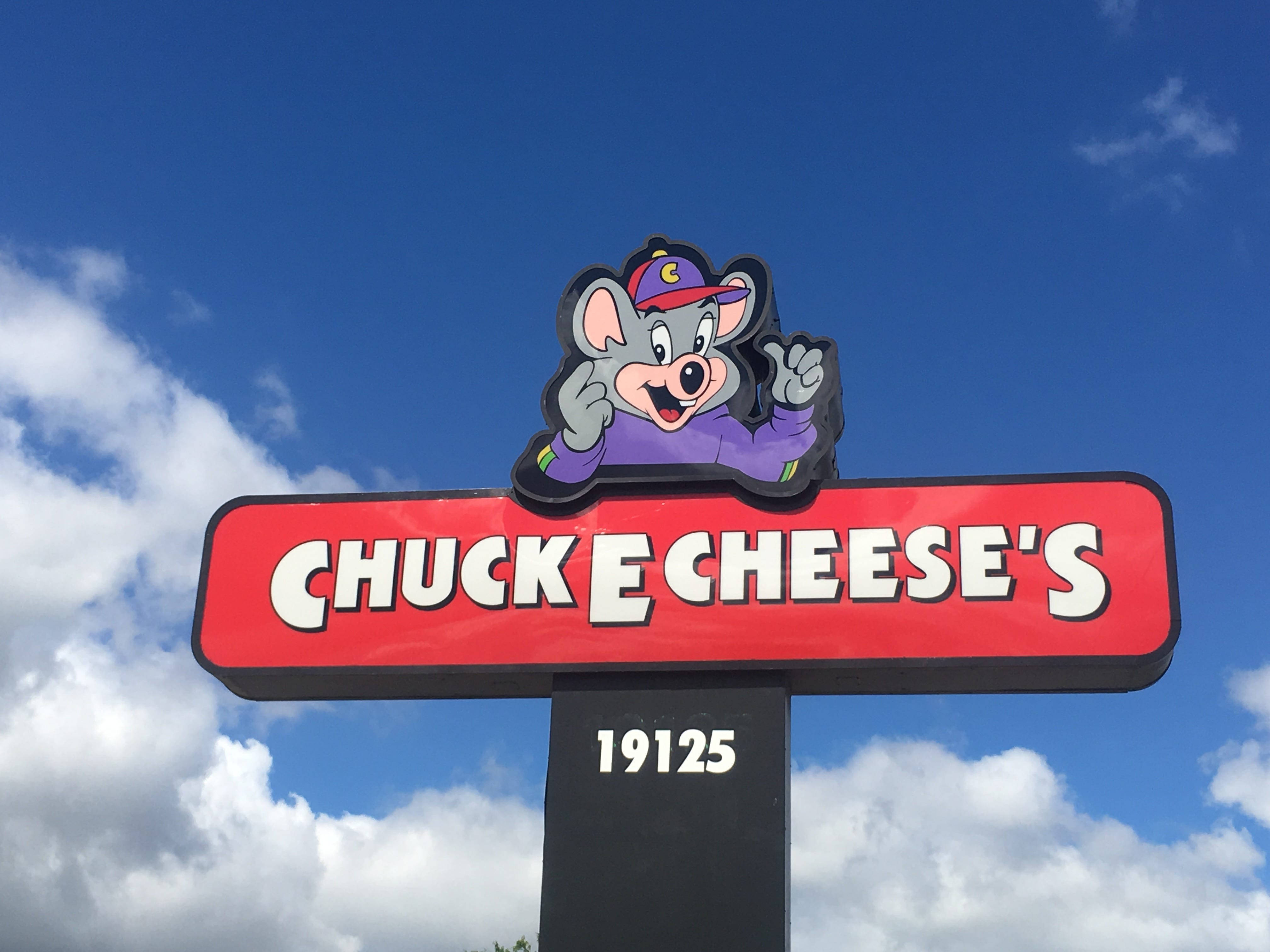 查克·E·切斯招牌 壁紙 — Chuck E Cheese 的吉祥物標誌，卡通老鼠在明亮的藍天和蓬鬆的白雲下揮手。