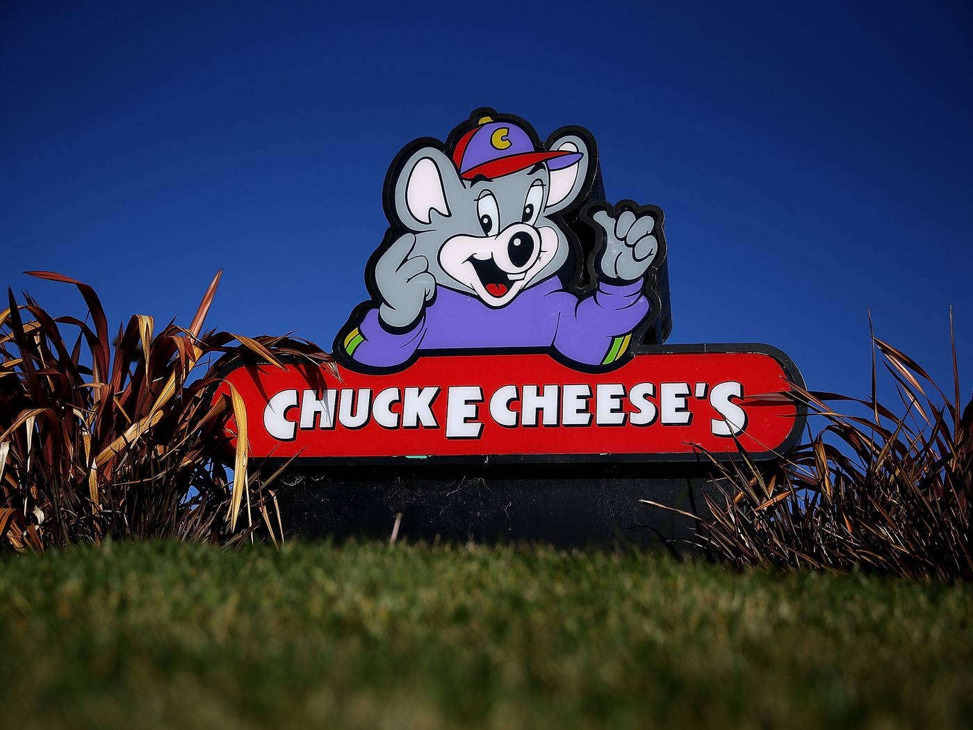 查克·E·切斯餐廳招牌 壁紙 — Chuck E Cheese 嘅吉祥物標誌，卡通老鼠揮手企喺草地上，背景係晴朗藍天。