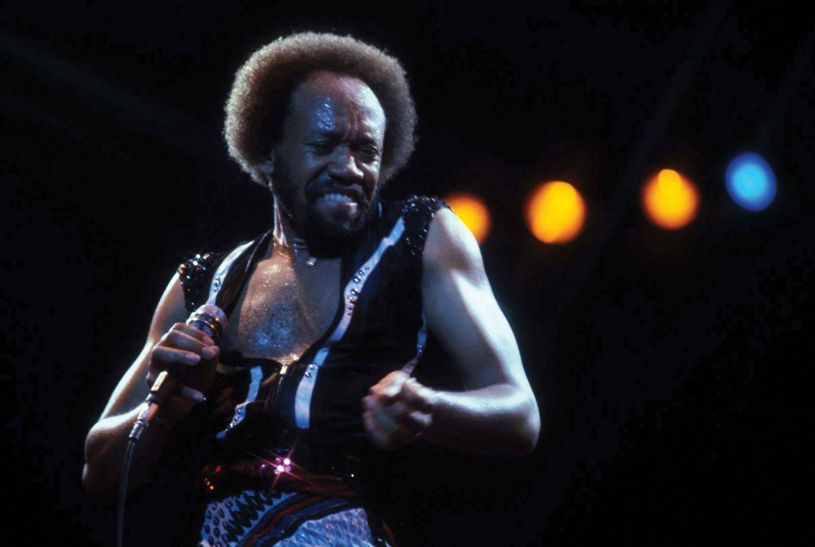 Earth, Wind And Fire 主唱 Maurice 持吉他姿勢 壁紙 — 一個汗流浹背的爆炸頭歌手在舞台燈光下對著咪高峰唱歌，背景漆黑中有發光的橙色和藍色散景。