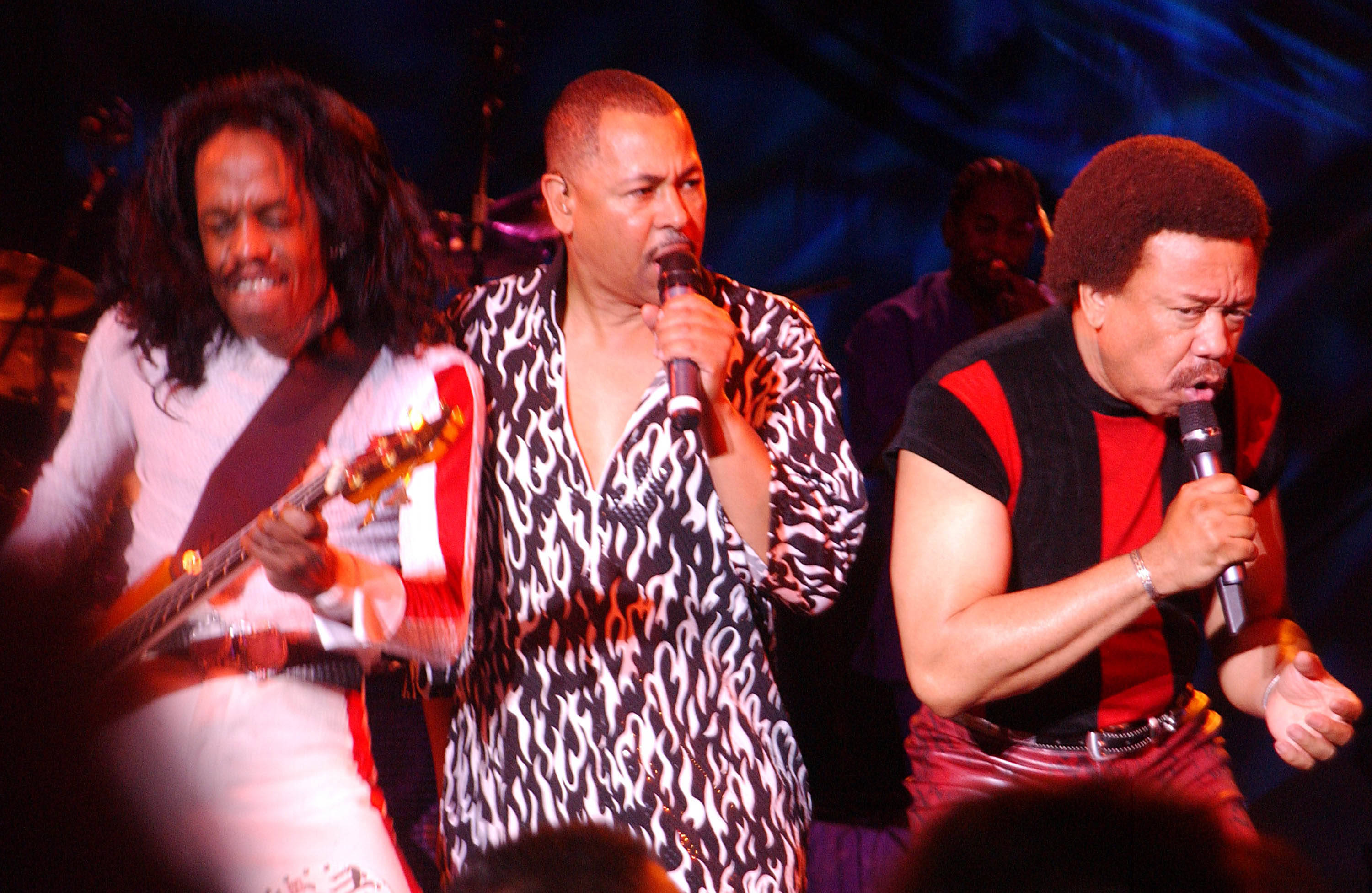 Earth, Wind And Fire 演唱《September》 壁紙 — 三位音樂家在充滿活力的舞台燈光下，帶著結他和麥克風充滿活力地表演。