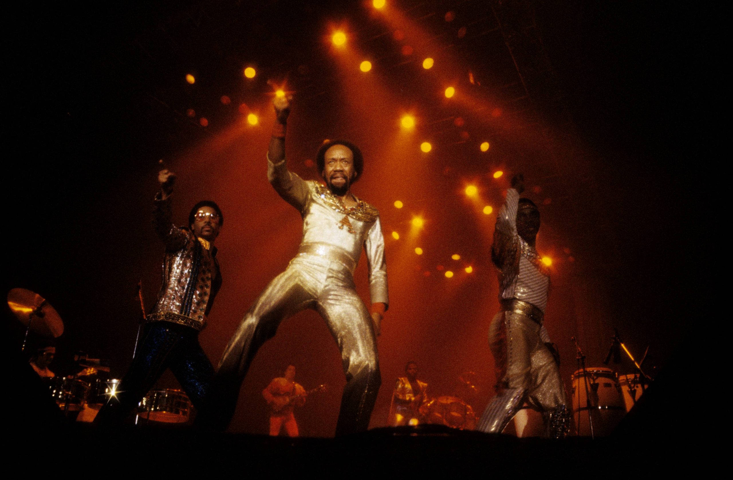 Earth, Wind And Fire 在倫敦溫布利現場演出 壁紙 — 樂隊在溫暖的橙色聚光燈下，穿著閃閃發光的服裝，以充滿活力的姿勢在舞台上表演。