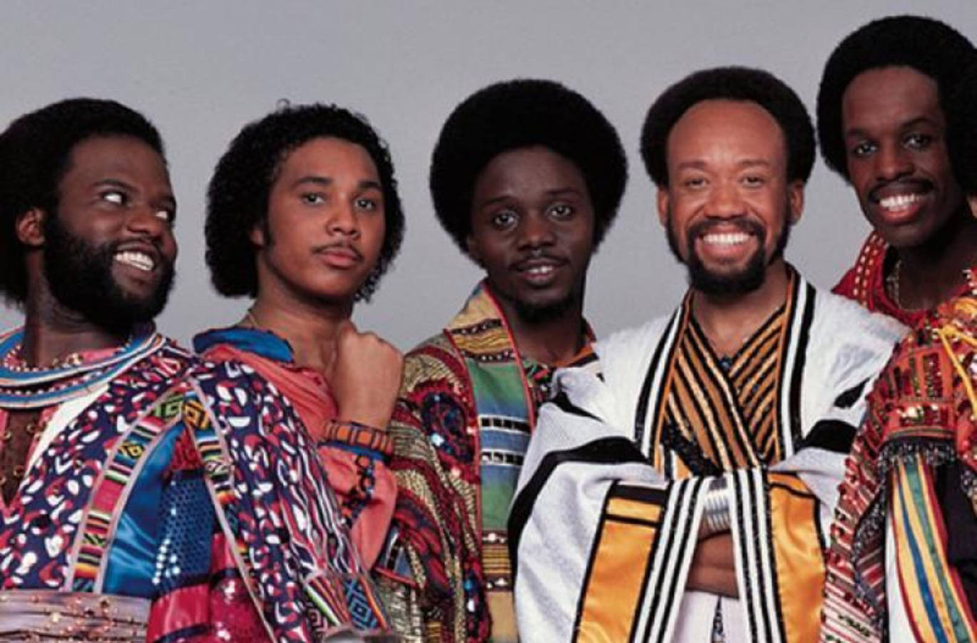 Earth, Wind And Fire 身著部落服裝 壁紙 — 五位微笑的黑人男性穿著鮮豔圖案的1970年代服裝，在純灰色工作室背景前自信地擺姿勢。