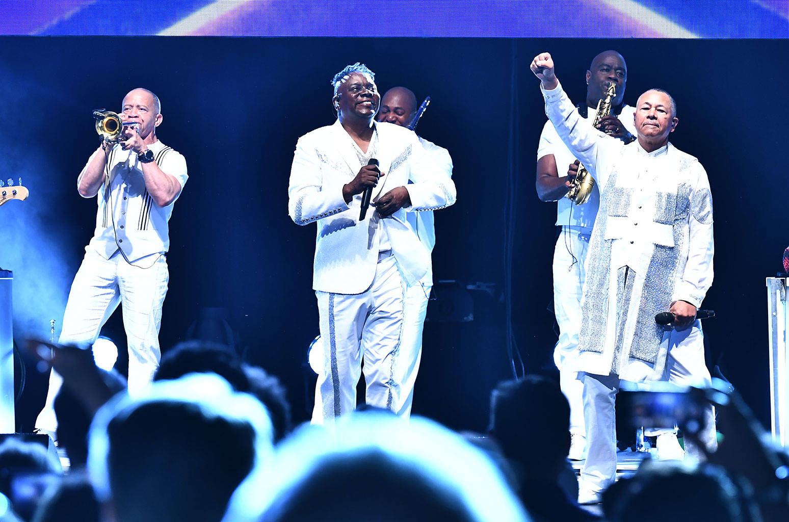 Earth, Wind And Fire 在年度晚宴演出 壁紙 — 一支身穿白色西裝的樂隊在藍色燈光下於舞台上充滿活力地表演，前景是觀眾的剪影。