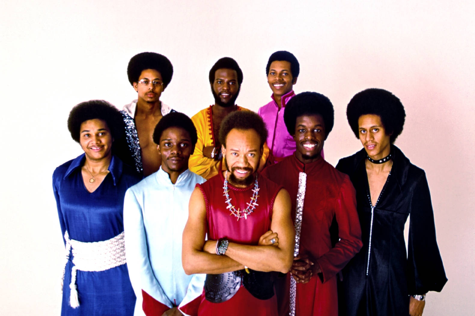 Earth, Wind & Fire 成員身穿繽紛絲質服裝 壁紙 — 七位留著非洲式髮型、穿著大膽70年代時尚的男士，在柔和的漸變背景前自信地擺姿勢。