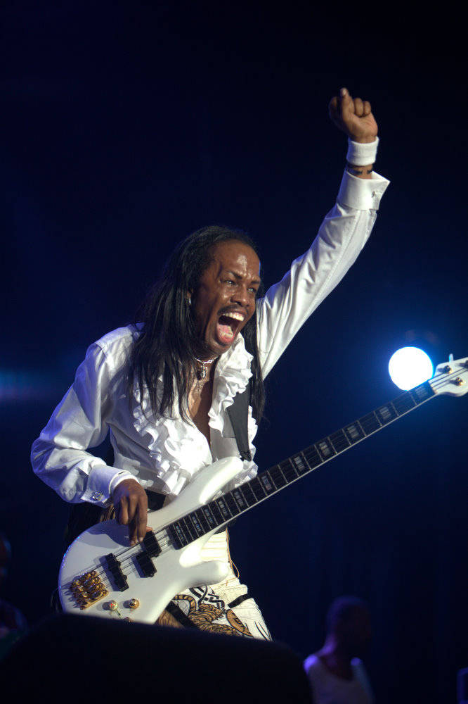 Earth, Wind & Fire 薇迪妮·懷特揮手致意 壁紙 — 一位留著長辮子的貝斯手在明亮的舞台燈光下，一邊彈奏白色貝斯吉他，一邊激情吶喊。