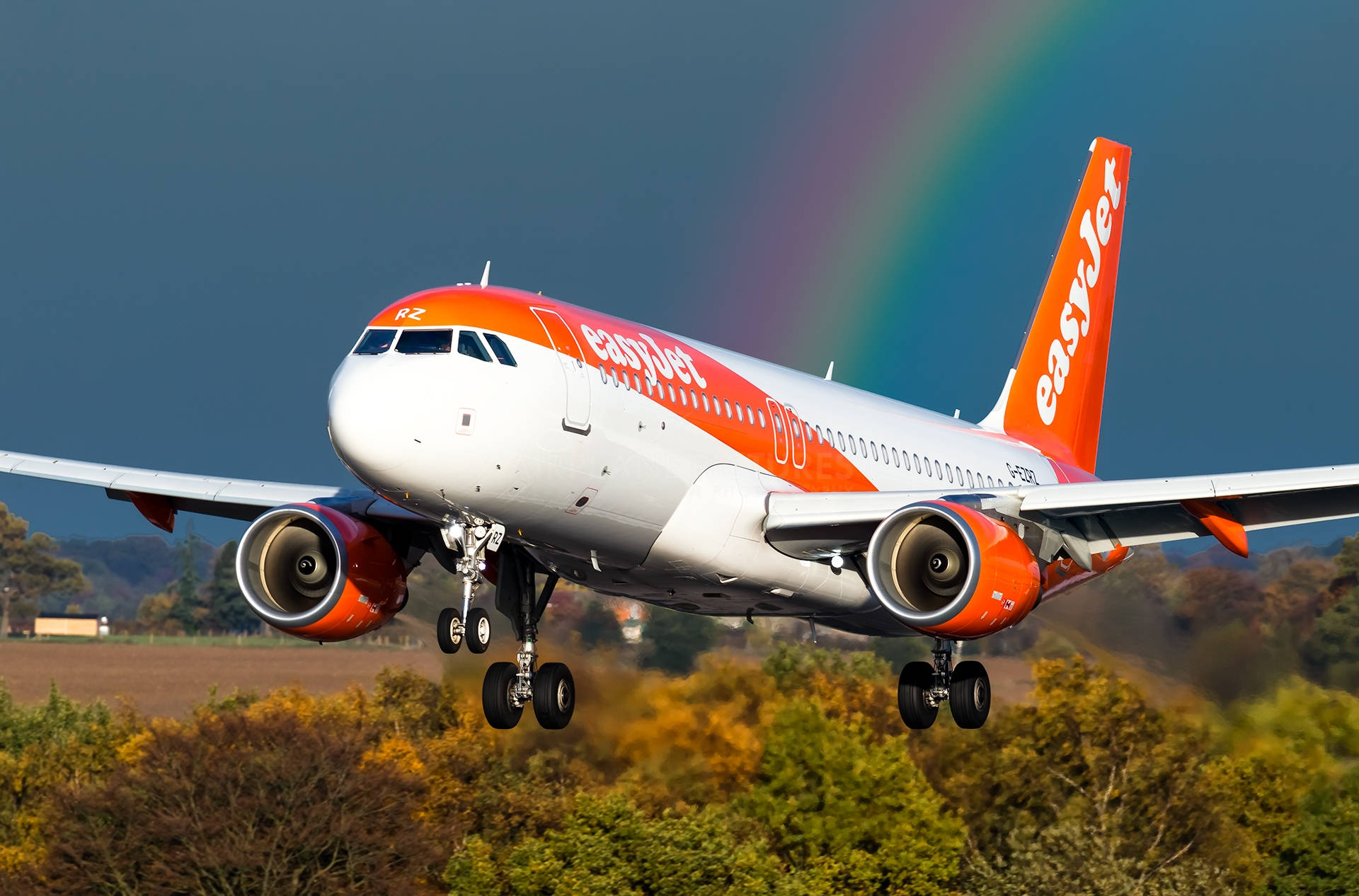 easyJet彩虹飛機 壁紙 — 一架易捷航空的飛機在戲劇性的天空下起飛，背後有一道鮮豔的彩虹拱起。