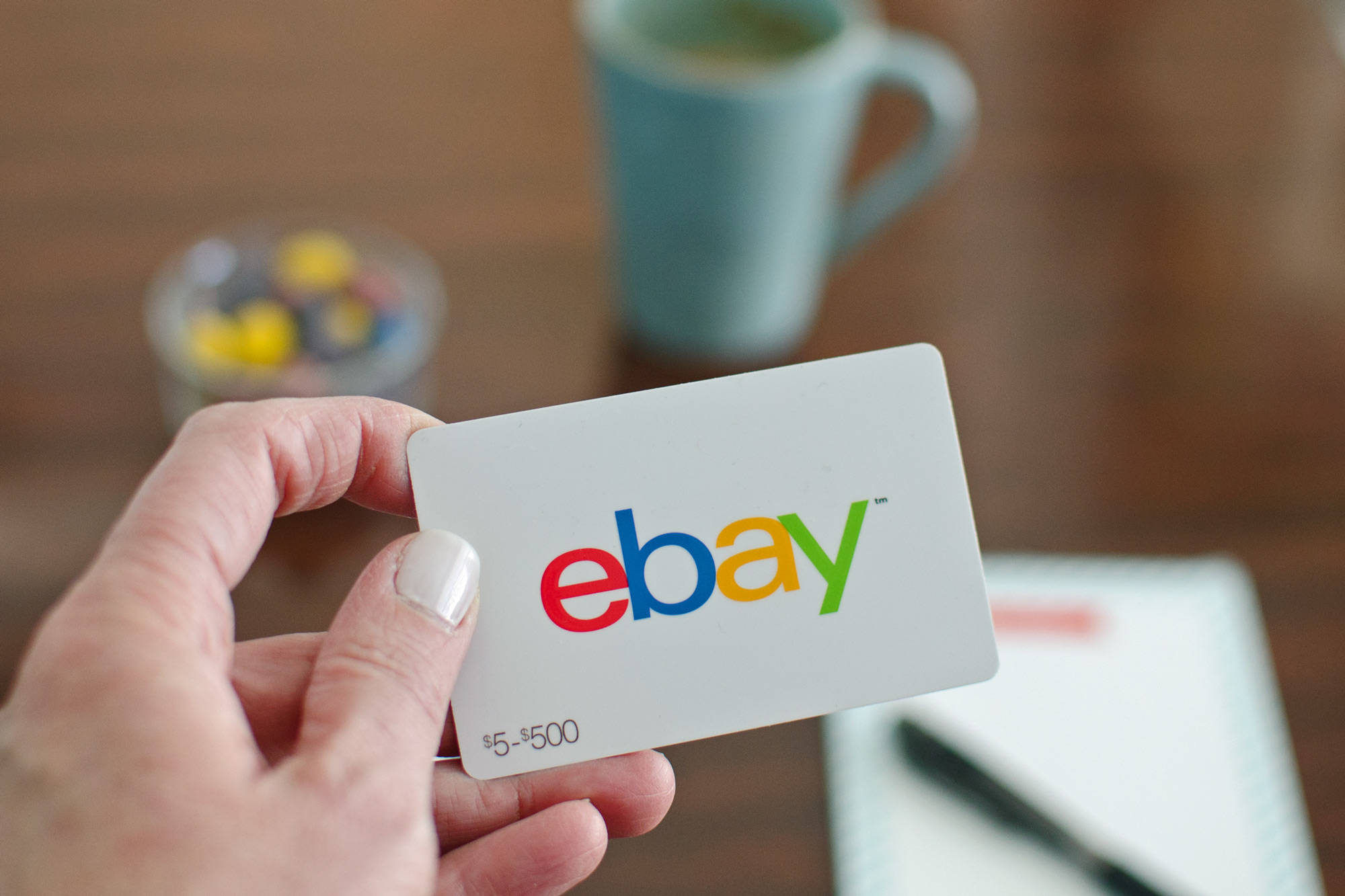 eBay 禮品卡 壁紙 — 一隻手拿著一張價值5至500美元的eBay禮品卡，背景是模糊的木桌，上面有杯子和糖果。