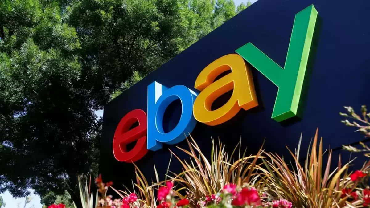 eBay英國標誌的低角度照片 壁紙 — 一個色彩繽紛的3D eBay標誌招牌在明亮的日光下，以綠樹和開花植物為背景，矗立在戶外。