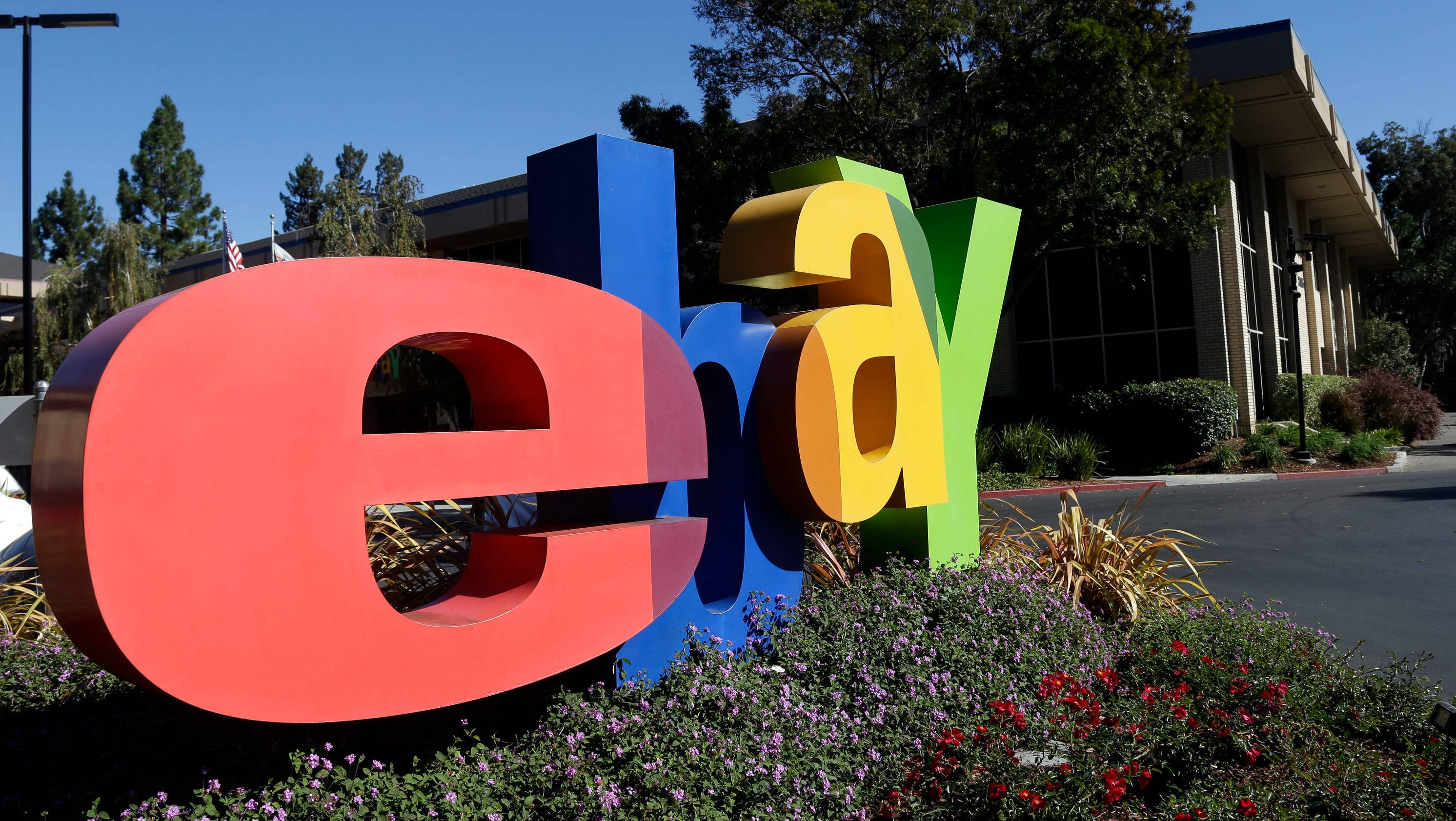 舊版 eBay 標誌 壁紙 — 在晴朗的藍天下，一座被開花灌木環繞的建築物前，放置著大型彩色eBay標誌字母。