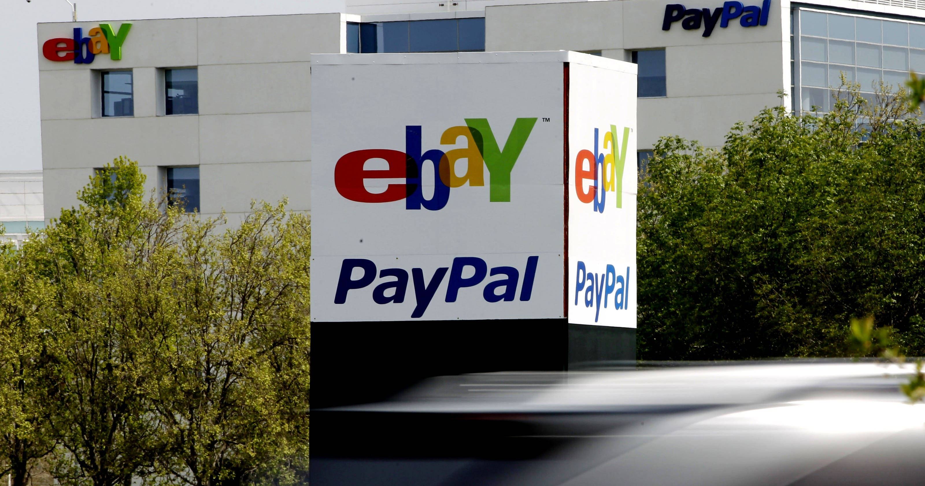 eBay 覆蓋 PayPal 壁紙 — 一個大型白色標牌展示色彩繽紛的eBay和藍色PayPal標誌，矗立在一棟現代辦公樓前，前景有綠色樹木。