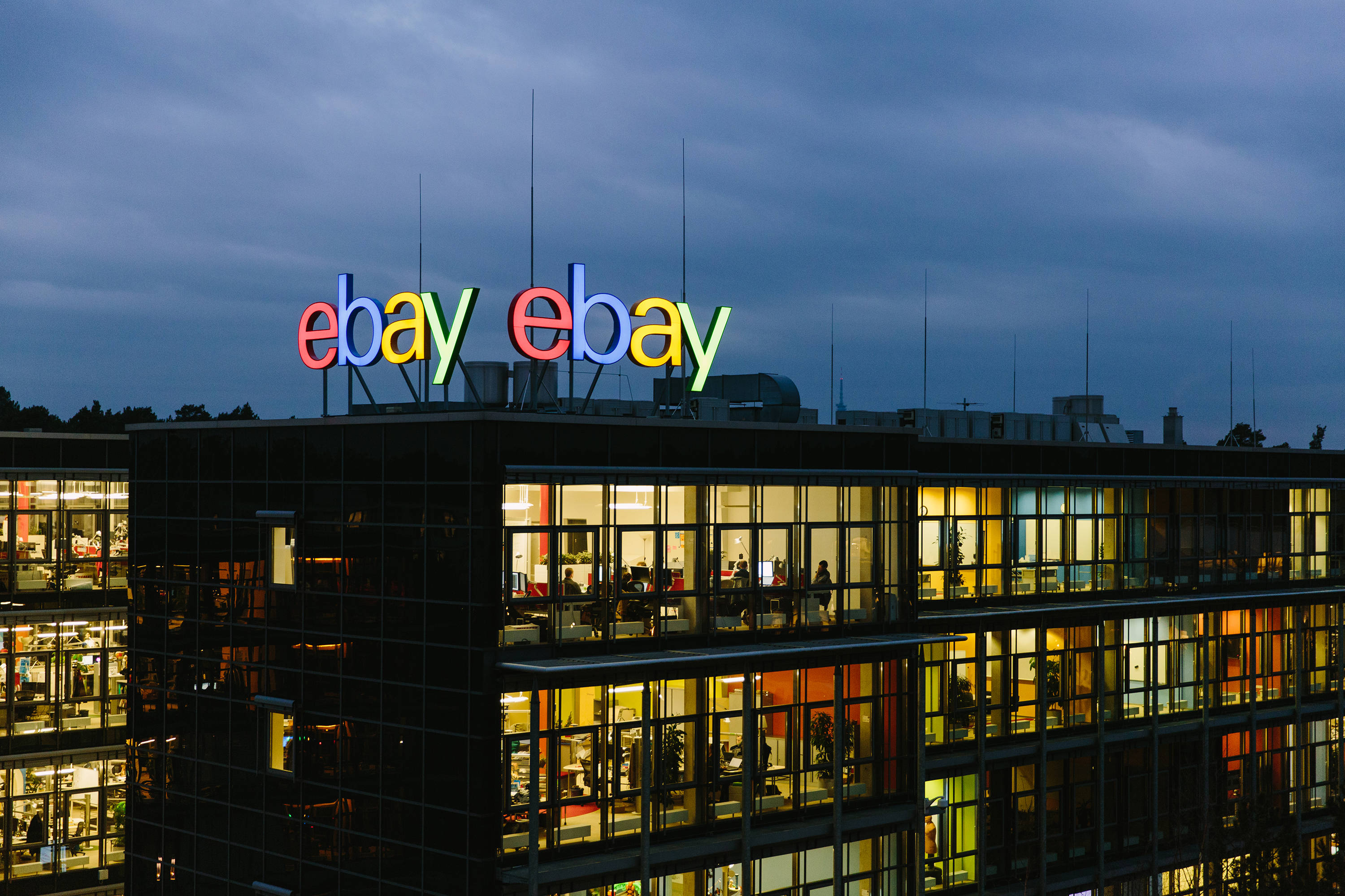 屋頂上的 eBay 標誌 壁紙 — 一座現代玻璃辦公大樓在黃昏時分，帶有發光的 'ebay' 標誌和溫暖的室內燈光，照亮內部的員工。