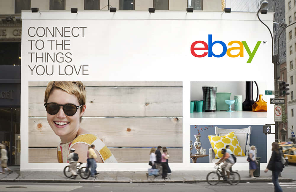 eBay UK 布幔 壁紙 — 城市街道上的一個大型eBay廣告牌展示了一位微笑的女性、色彩繽紛的家居裝飾和臥室場景，行人和騎自行車的人經過。