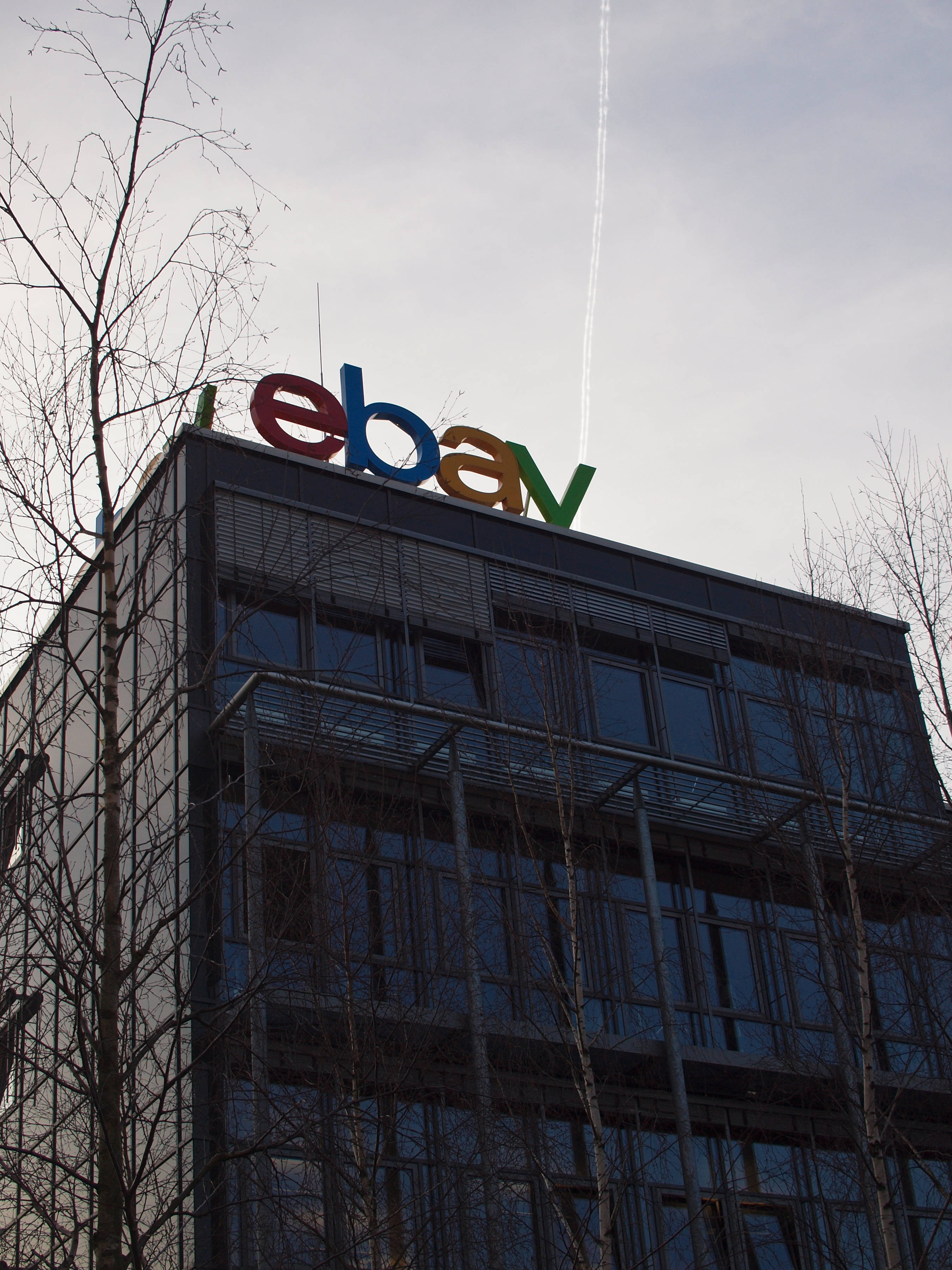 eBay 標誌高樓 壁紙 — 一座現代玻璃建築，頂部有色彩繽紛的eBay標誌，襯著蒼白的天空，光禿禿的樹木框住畫面。