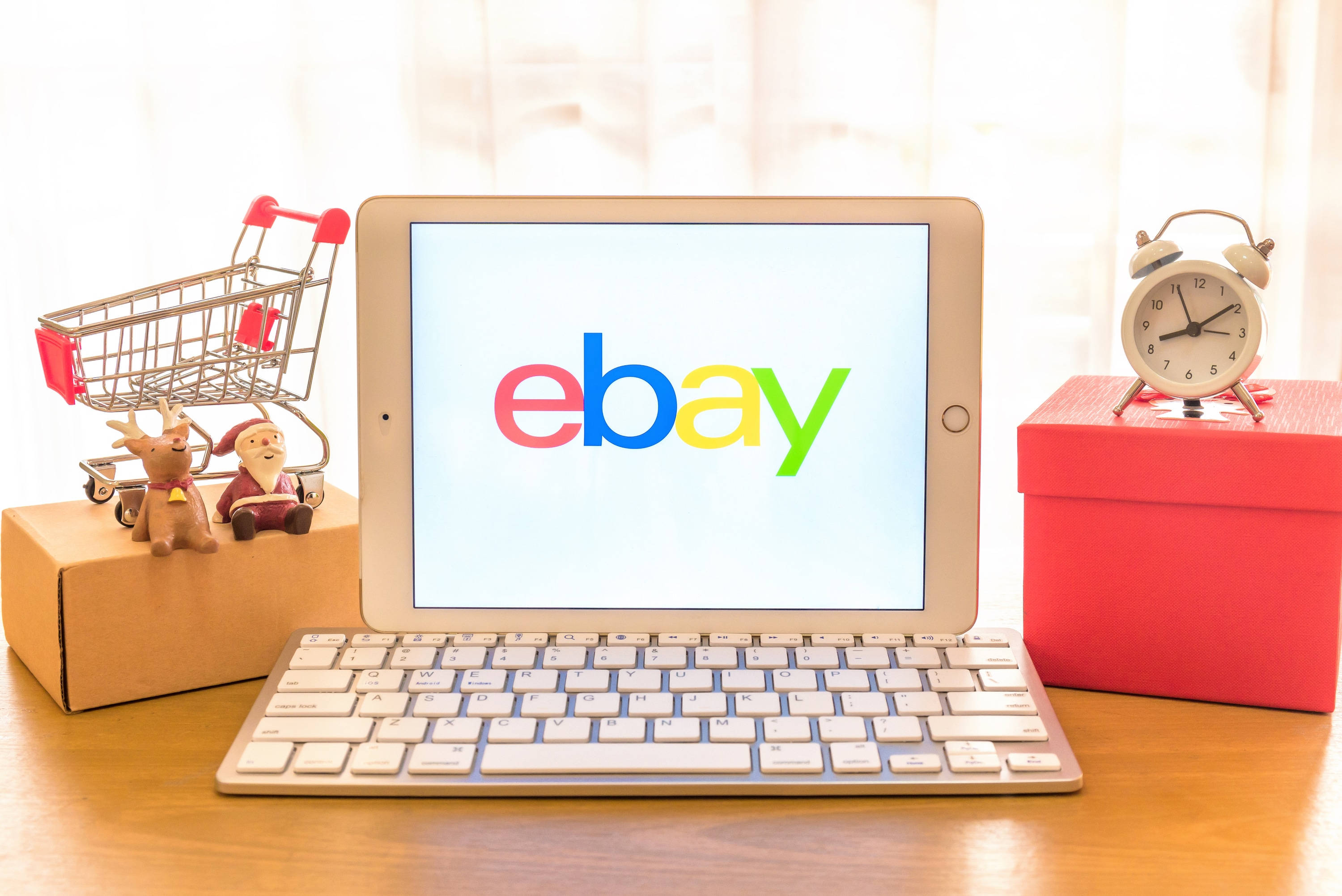 eBay 平板電腦 壁紙 — 一部顯示eBay標誌的平板電腦放在木桌上，旁邊有一個微型購物車、聖誕老人公仔和一個帶鬧鐘的紅色禮物盒。