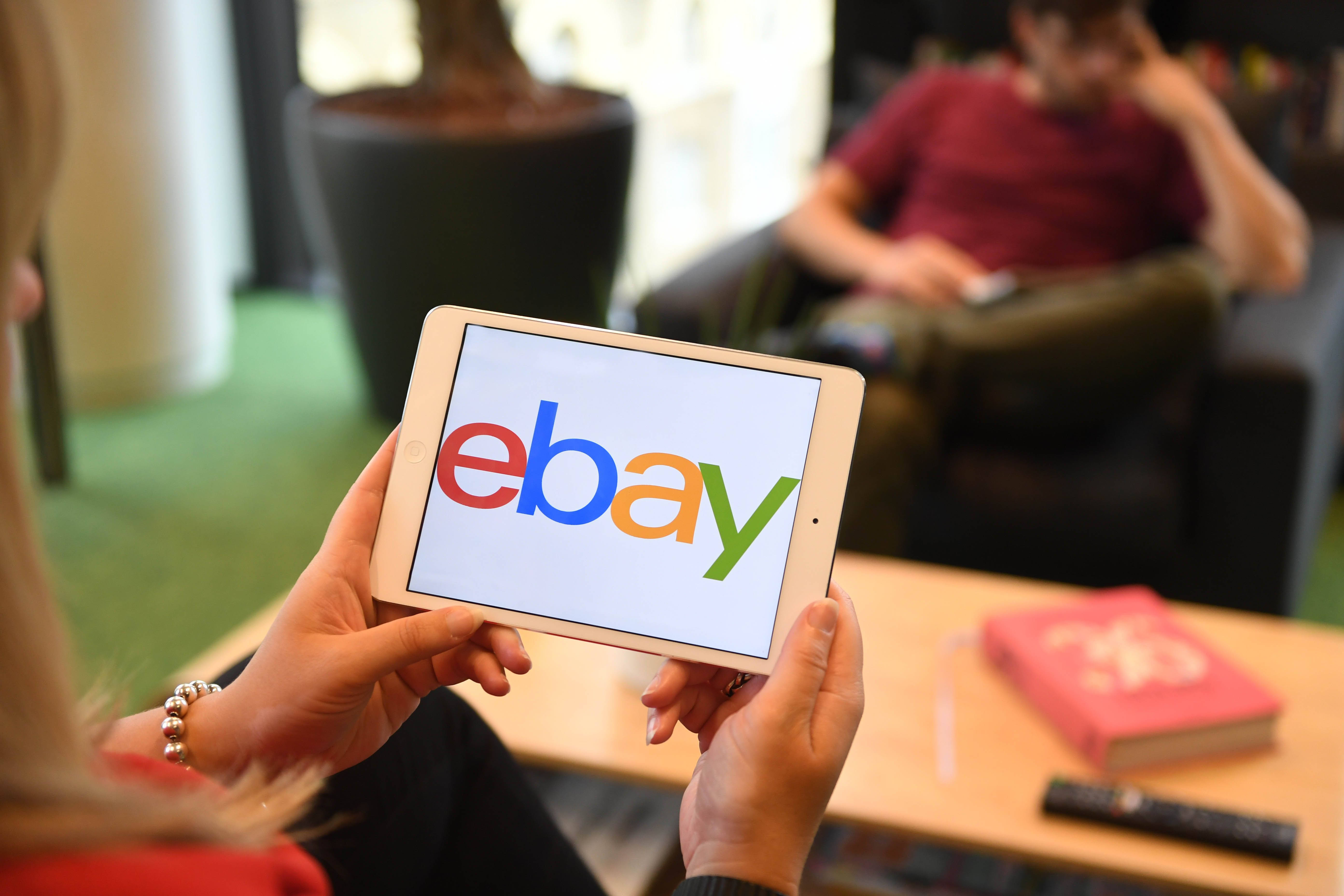 eBay 標誌平板電腦 壁紙 — 一個人坐在現代客廳中，手持顯示eBay標誌的平板電腦，背景有另一個人。