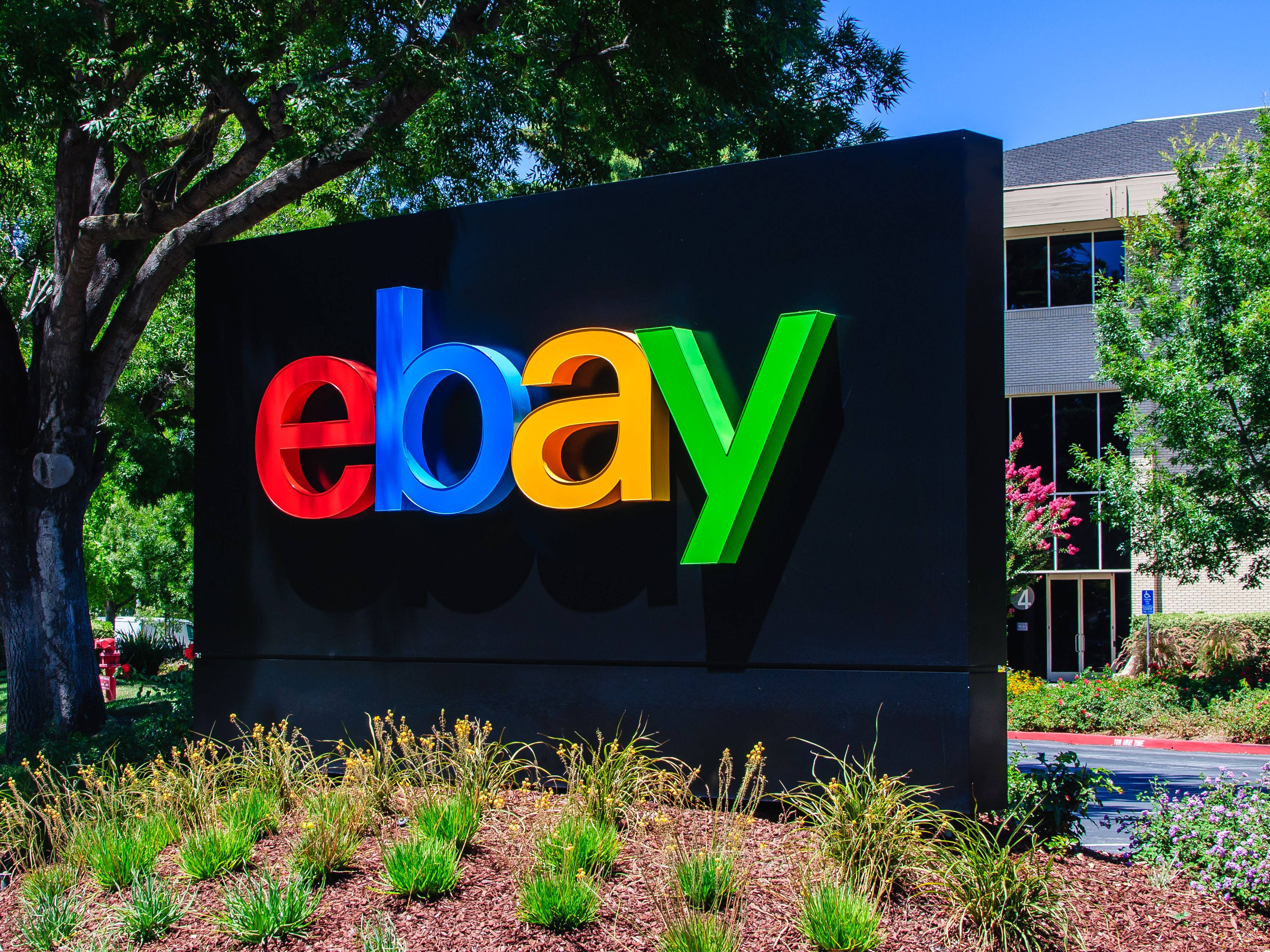 eBay 黑色標誌 壁紙 — 一個鮮豔的eBay標誌招牌，帶有紅色、藍色、黃色和綠色的字母，矗立在戶外，在晴朗的藍天下，周圍環繞著茂密的綠樹和園林植物。