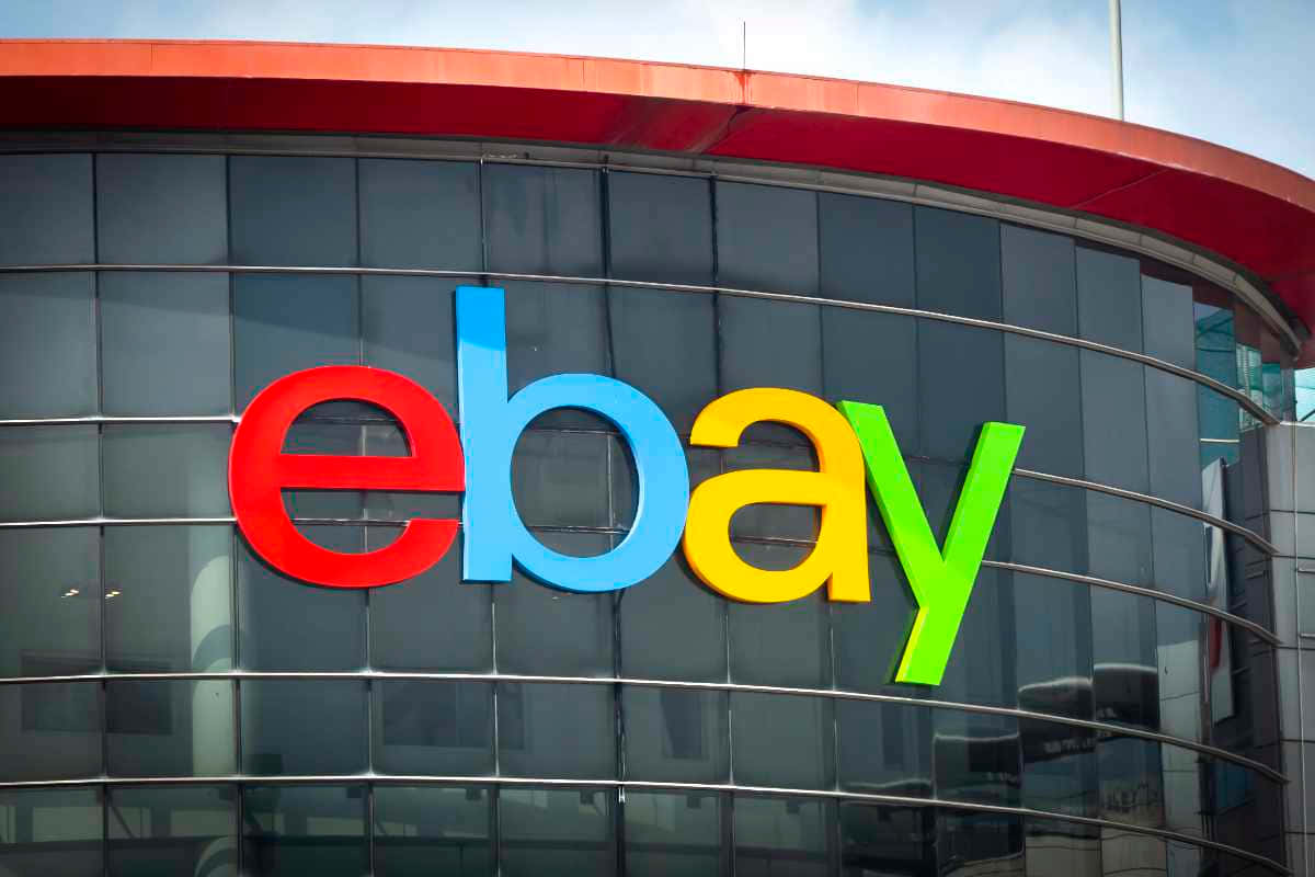 eBay英國標誌於玻璃窗上 壁紙 — 色彩繽紛的eBay標誌，以粗體紅、藍、黃、綠色字母組成，安裝在現代玻璃建築外牆上，天空部分多雲。