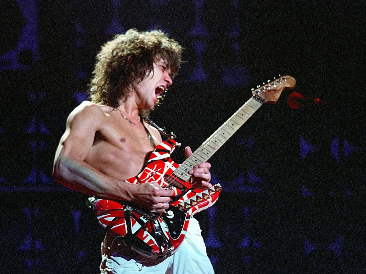 Eddie Van Halen Frankenstrat 吉他演奏 壁紙 — 一位赤裸上身的搖滾結他手，頭髮狂野，在黑暗的舞台上熱情地尖叫，同時彈奏一把紅白電結他。