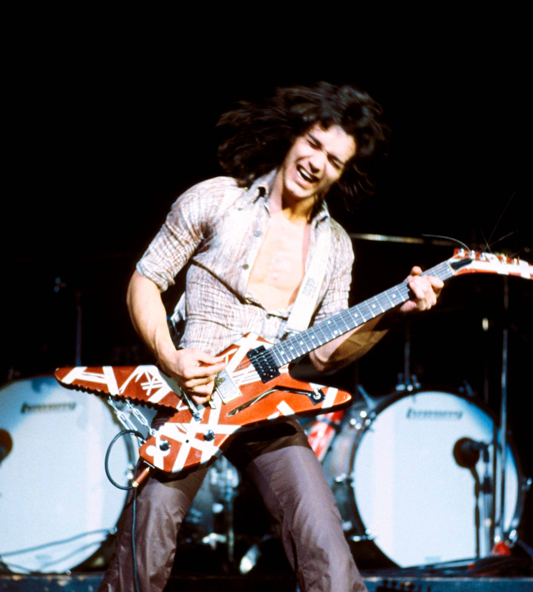 Eddie Van Halen 搖滾音樂 壁紙 — 一位充滿活力的搖滾結他手，頭髮狂野，在明亮的燈光下於舞台上彈奏紅白條紋電結他。
