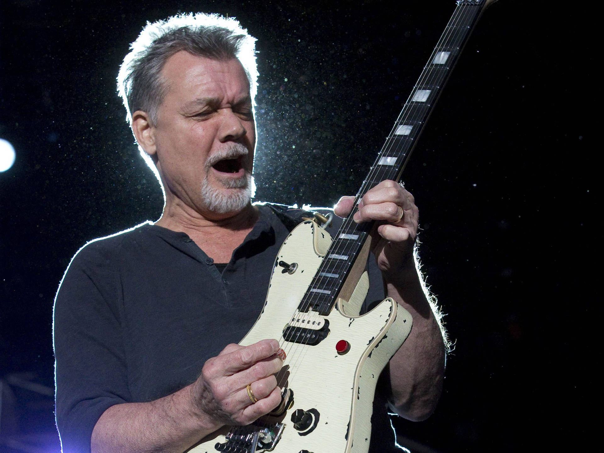 Eddie Van Halen 搖滾吉他手 壁紙 — 一位留著白鬍子的中年男子在明亮的舞台燈光下熱情地彈奏一把破舊的奶油色電結他。