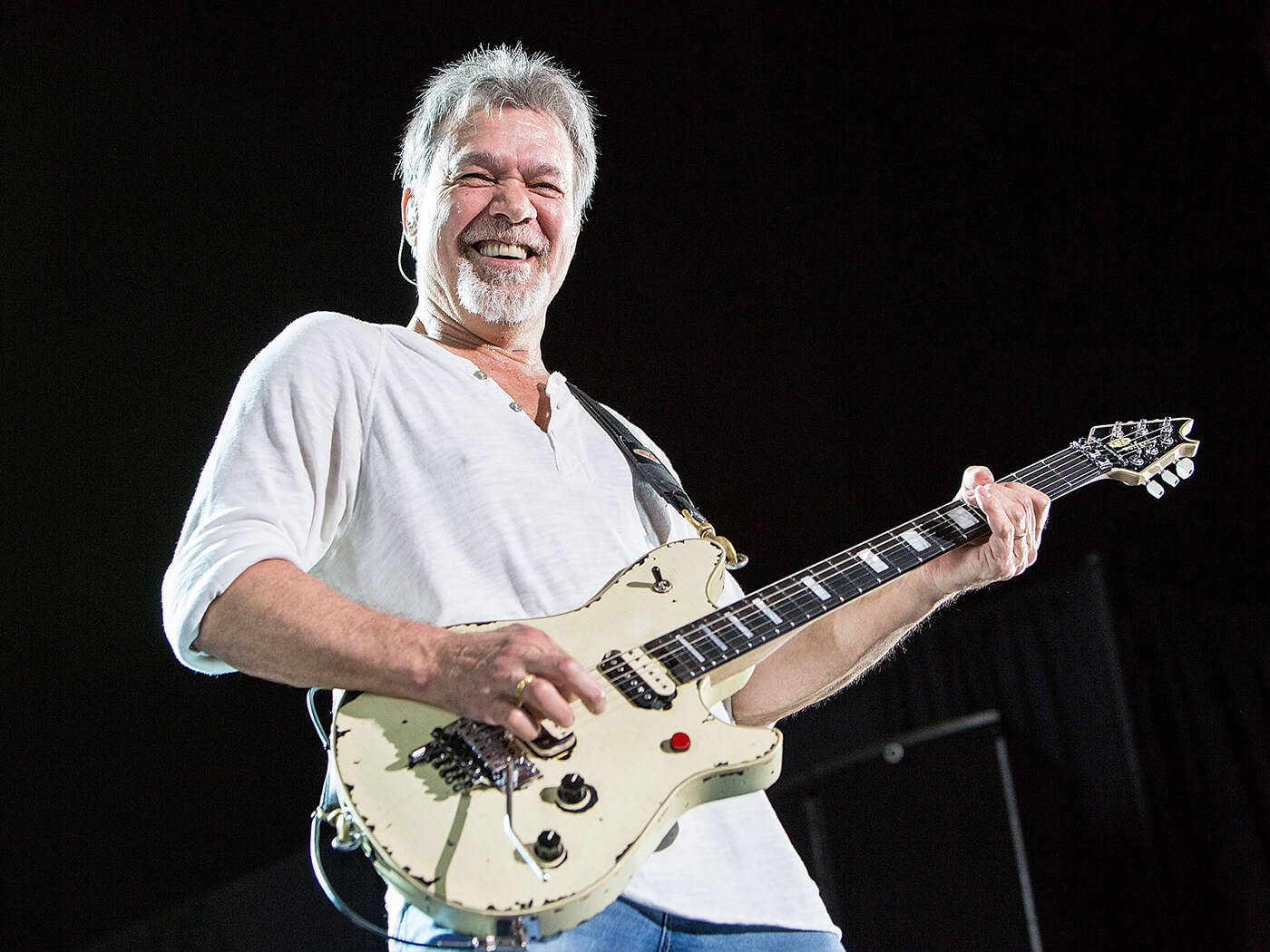 Eddie Van Halen 搖滾電吉他 壁紙 — 一位快樂的灰髮音樂家在黑暗的舞台上彈奏奶油色電結他，散發光芒。