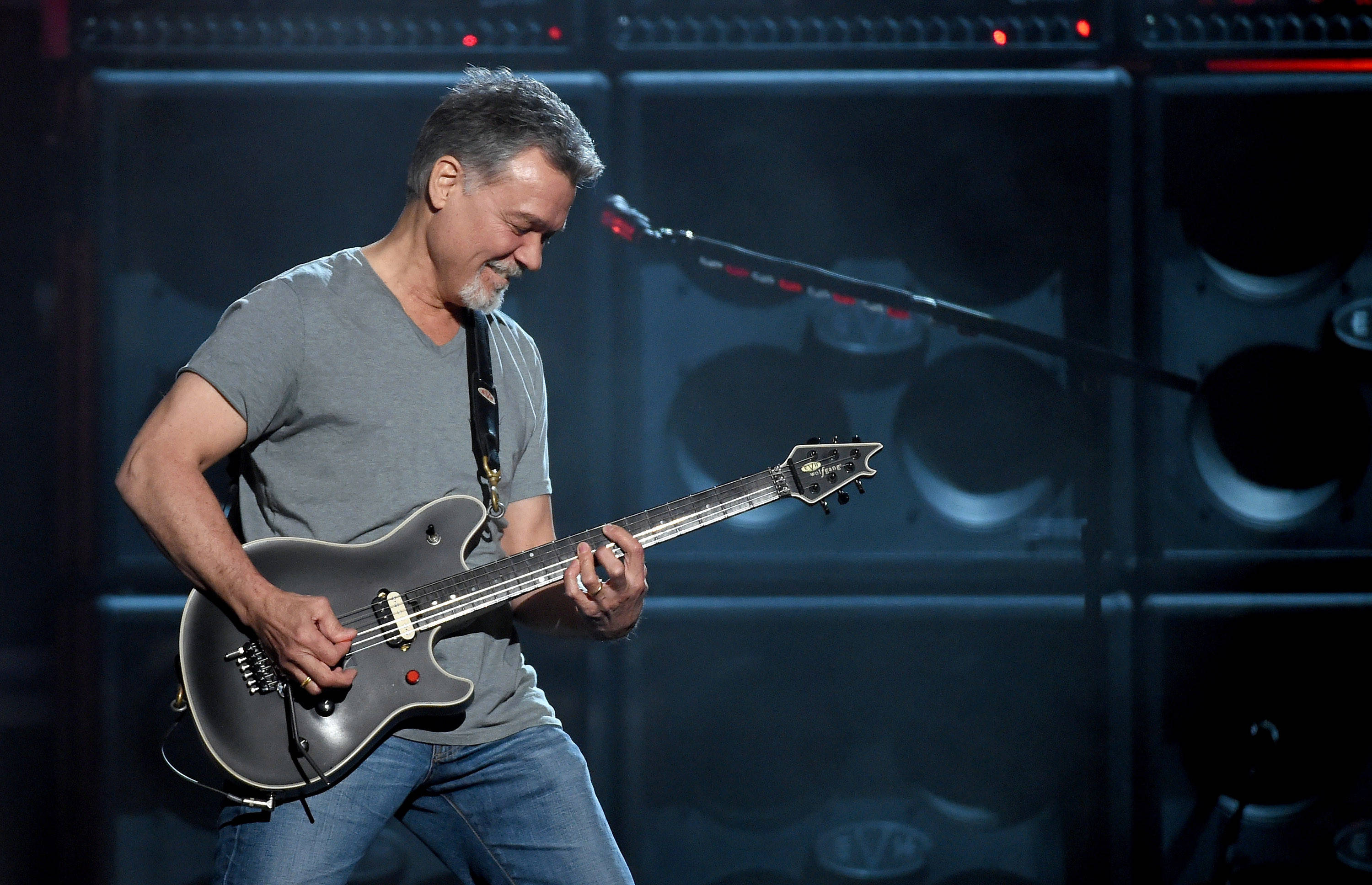 Eddie Van Halen 樂隊 演唱會巡迴 壁紙 — 一位留鬍鬚的中年男子在燈光昏暗的舞台上熱情地彈奏黑色電結他，身後有大型揚聲器。