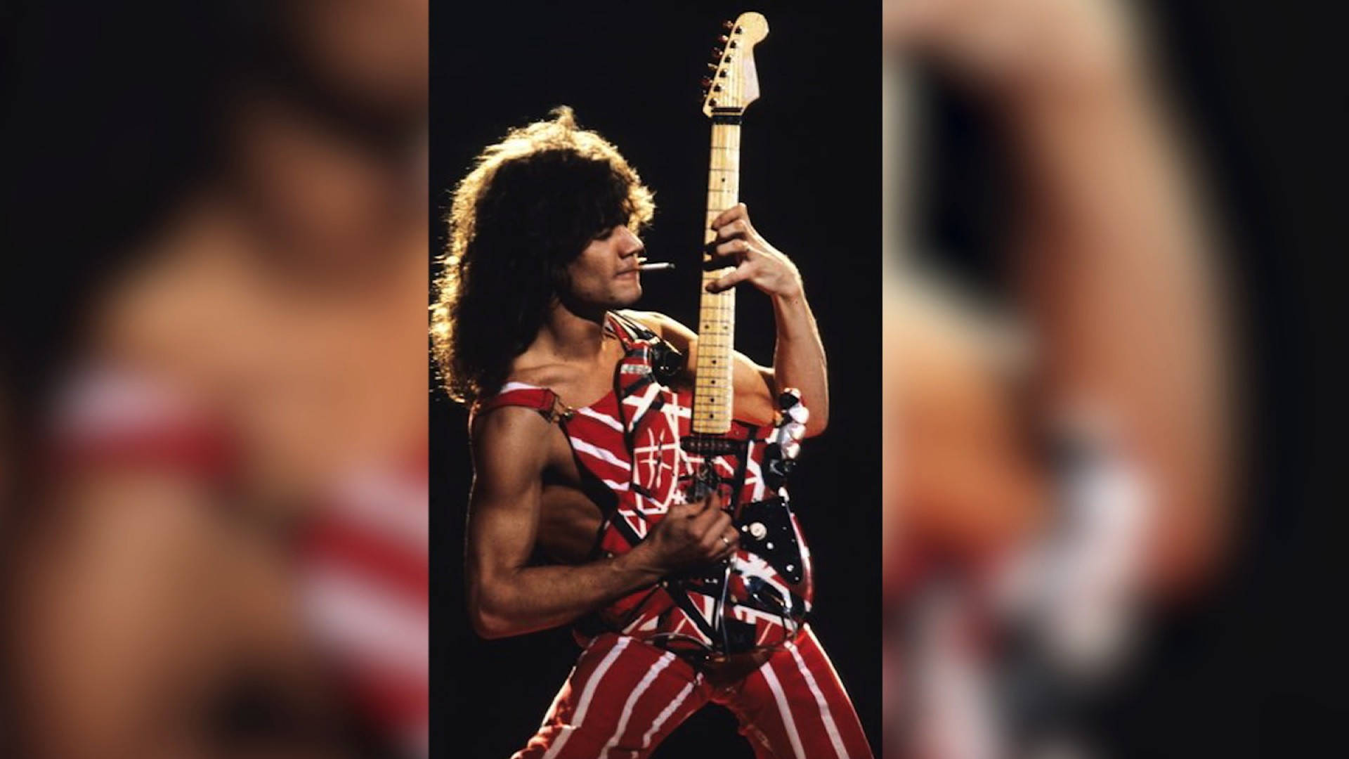 Eddie Van Halen 樂隊吉他演奏 壁紙 — 一位捲髮搖滾結他手，身穿紅白條紋服裝，在黑暗舞台背景下抽煙，彈奏 Fender Stratocaster。