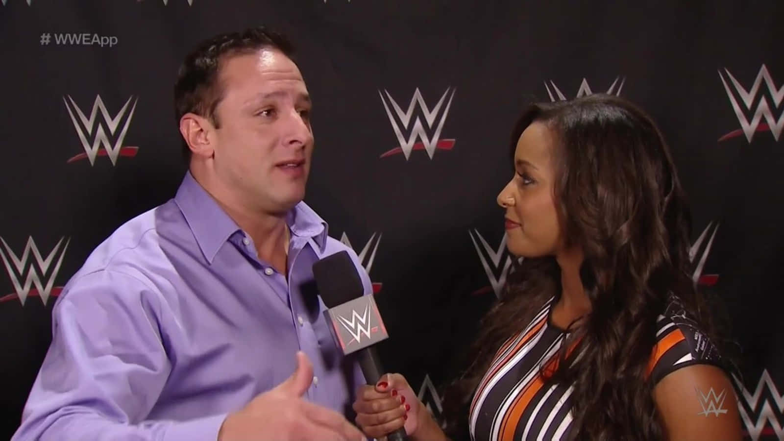 Eden 與摔角手 Billy Kidman 對談 壁紙 — 一位穿著紫色襯衫的WWE採訪者對著一位女採訪者手持的麥克風說話，背景為黑色WWE標誌。