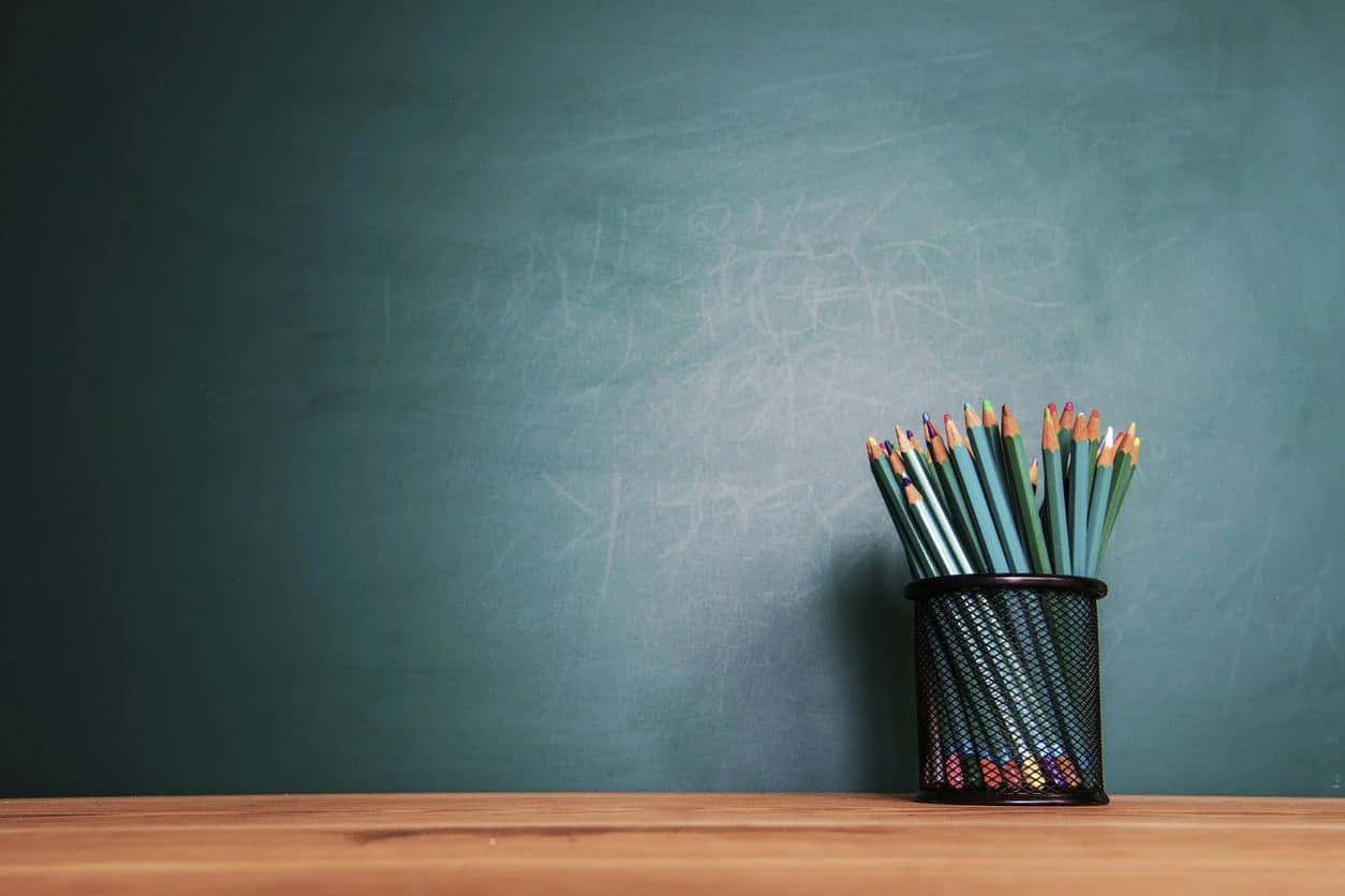彩色鉛筆放在黑色網狀筆筒中，置於木桌上，前面是綠色黑板。